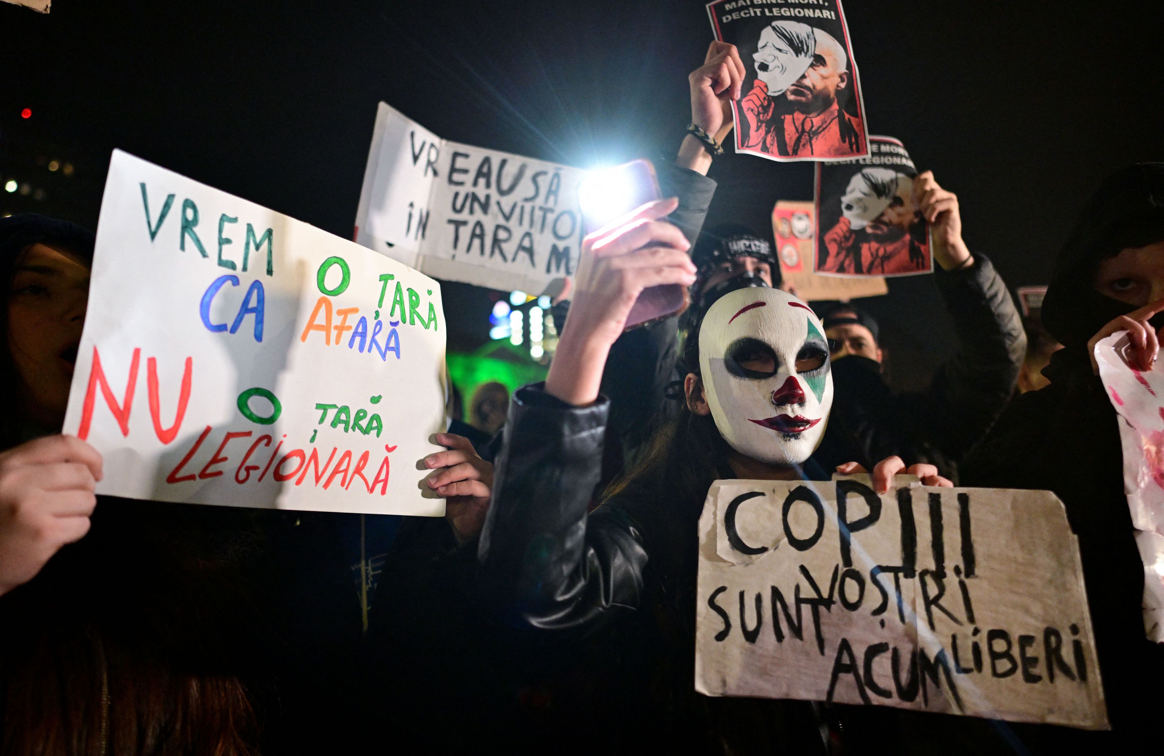 Ludzie protestują trzymając kartony z napisami po rumuńsku. Na pierwszym planie człowiek w masce arlekina