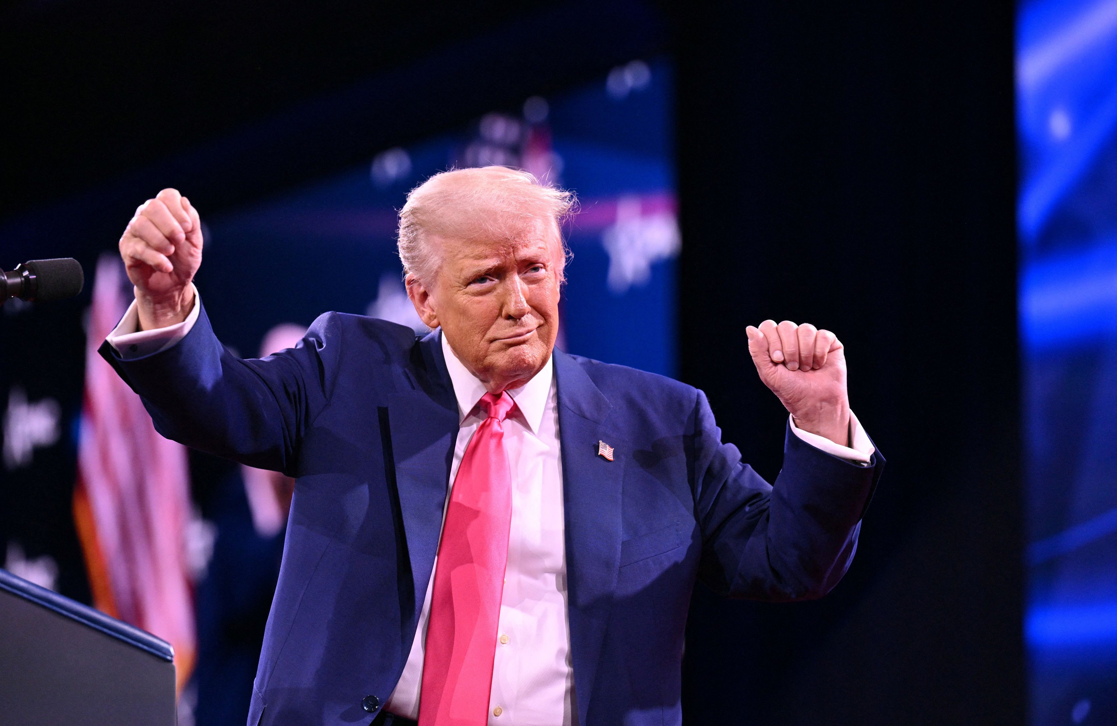 Starszy mężczyzna w niebieskim garniturze i różowym krawacie (prezydent USA Donald Trump) tańczy na scenie z uniesionymi w górę rękami