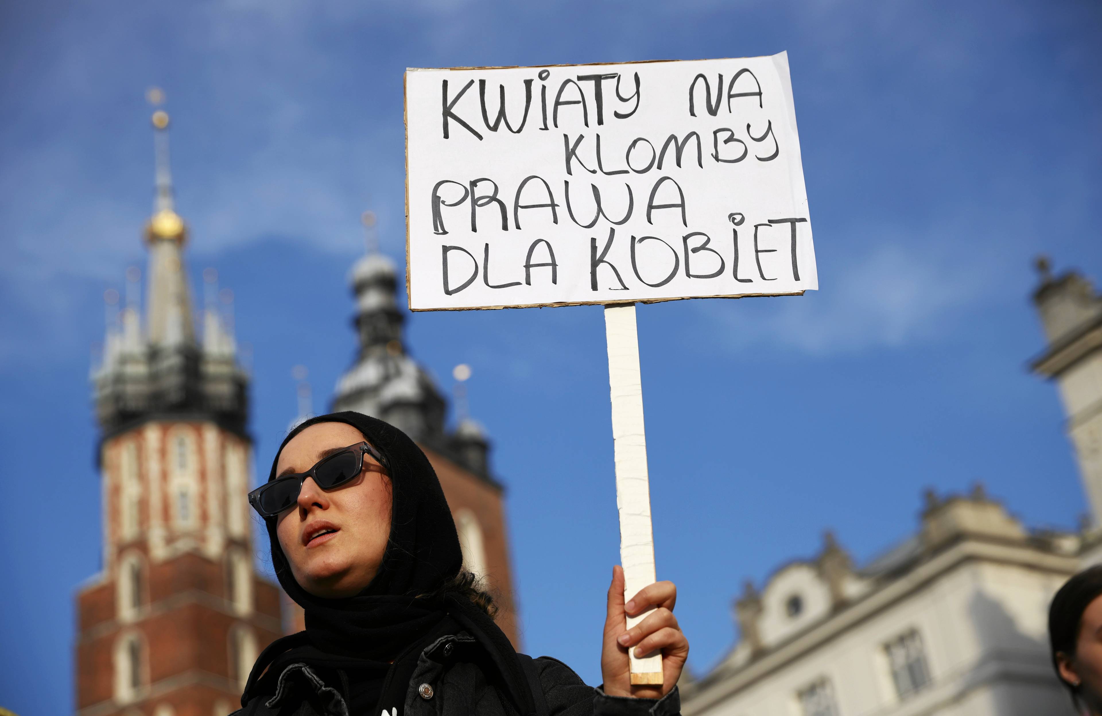 kobieta w ciemnych okularach i czarnej chustce trzyma transparent z napisem „Kwiaty na klomb, prawa dla kobiet". W tle dwie nierówne wieże Kościoła Mariackiego