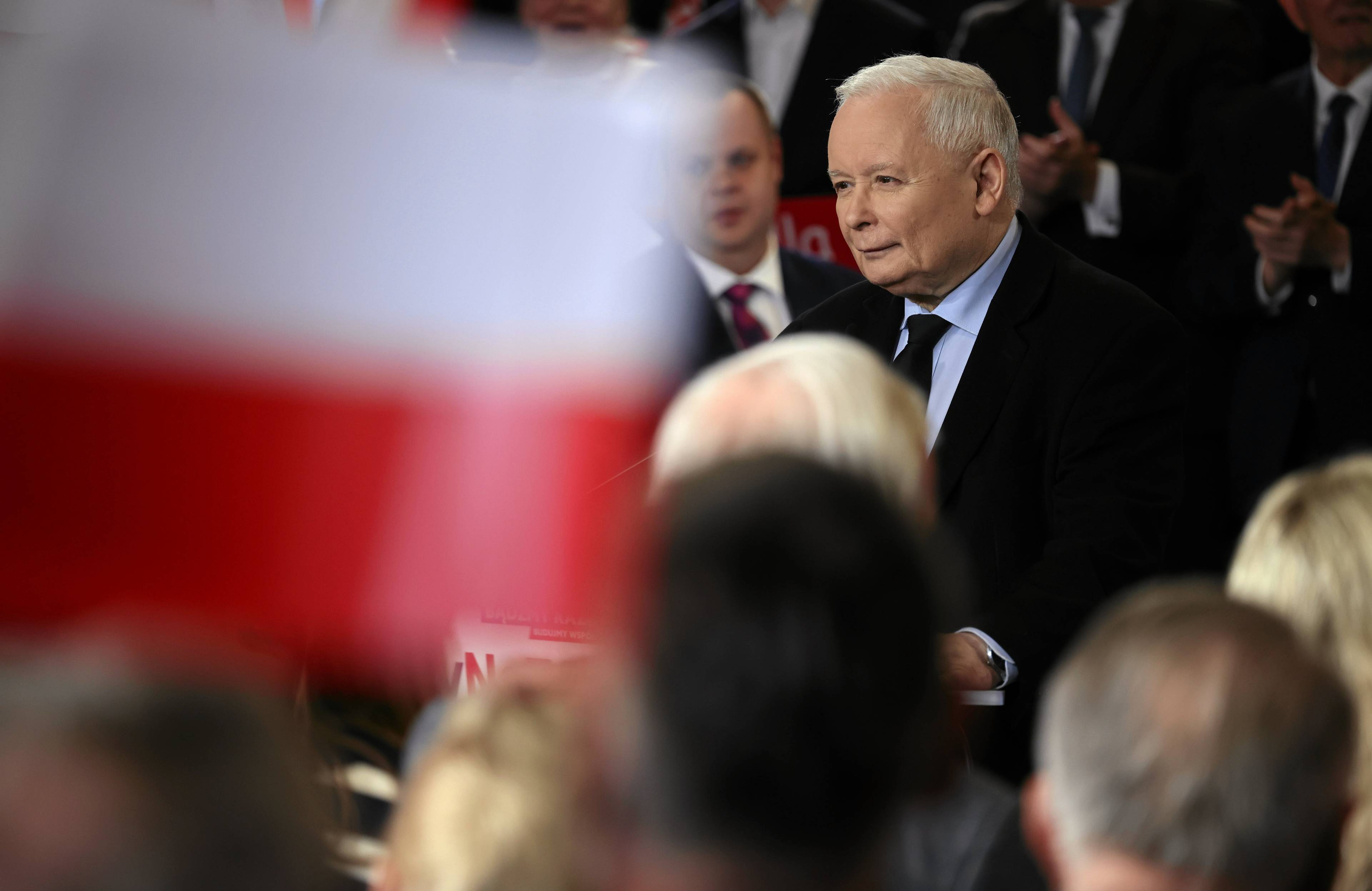 Jarosław Kaczyński w garniturze wśród ludzi na wiecu wyborczym, na pierwszym planie obok polityka lekko rozmyta flaga Polski