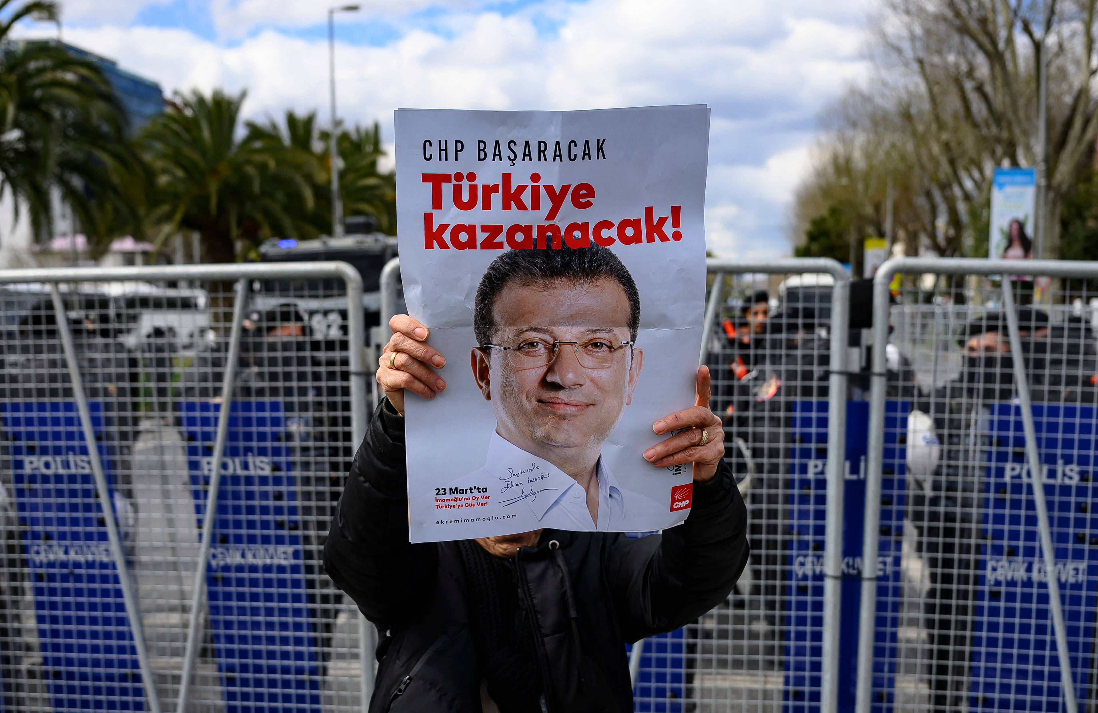 Protestujący trzyma plakat z wizerunkiem Ekrema Imamoglu, zatrzymanego burmistrza Stambułu, w tle blokada policyjna