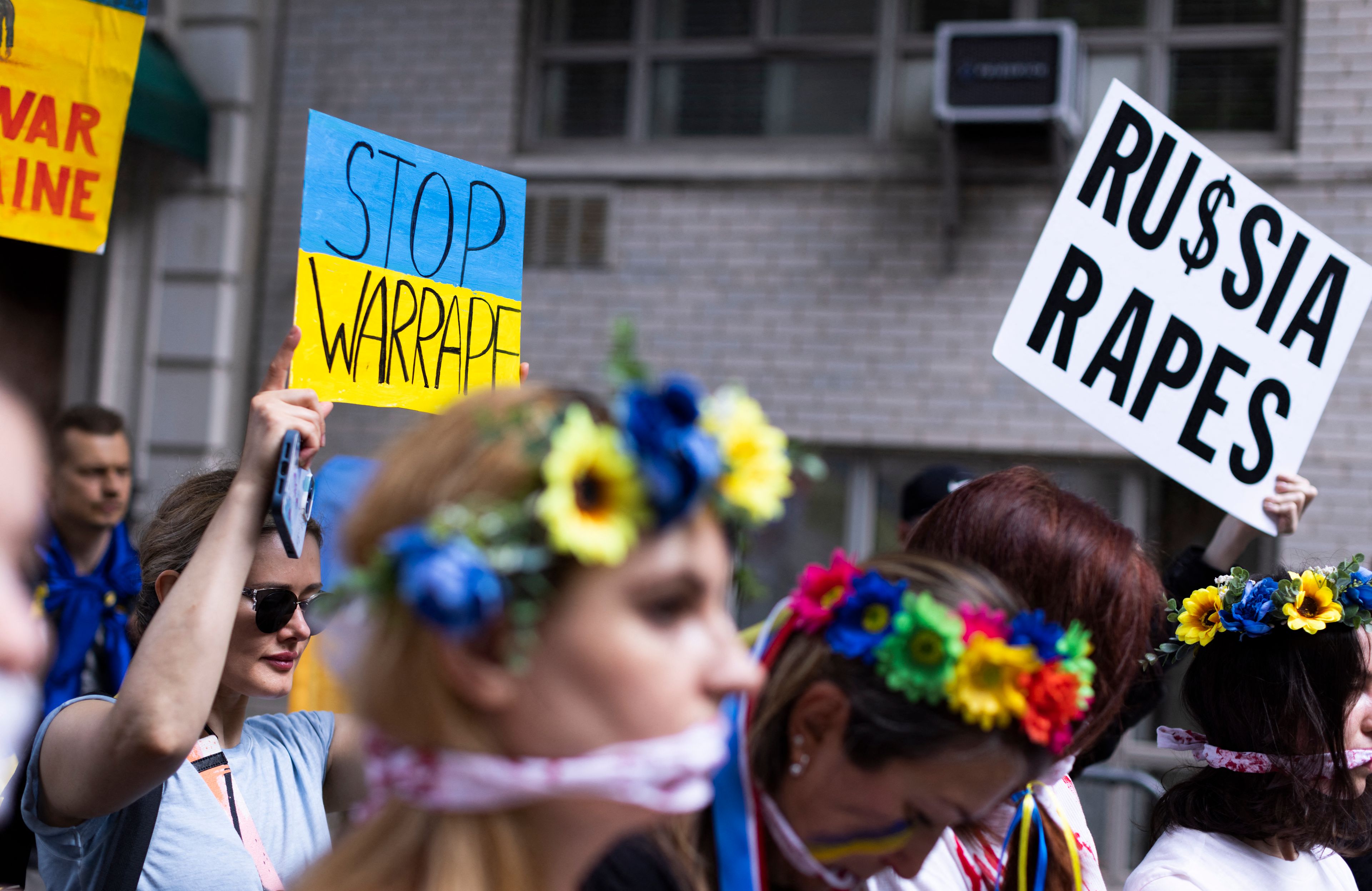 Demonstracja przeciwko rosyjskiej agresji na Ukrainę, na pierwszym planie dzieczyna w niebiesko-żółtym wianku na głowie, w tle plakat Russia Rapes - Rosja gwałci. Przemoc seksualna na wojnie