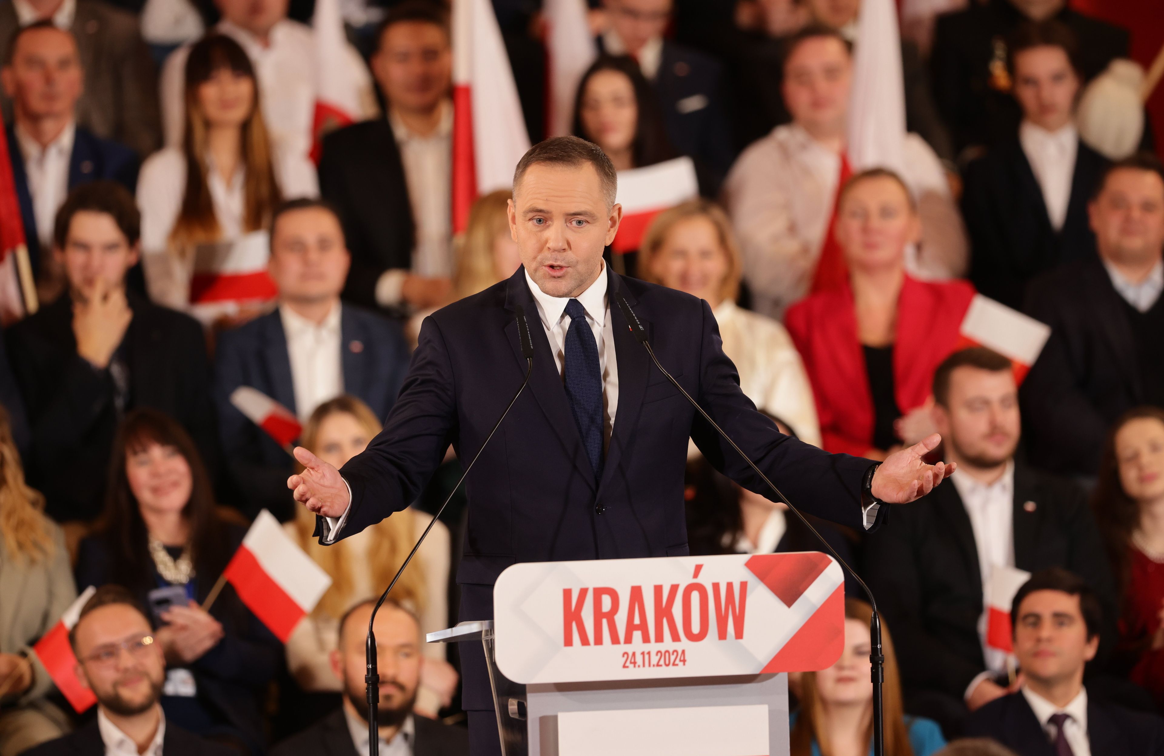 40-latek w garniturze na podium, za nim ludzie z biało-czerwonymi flagami