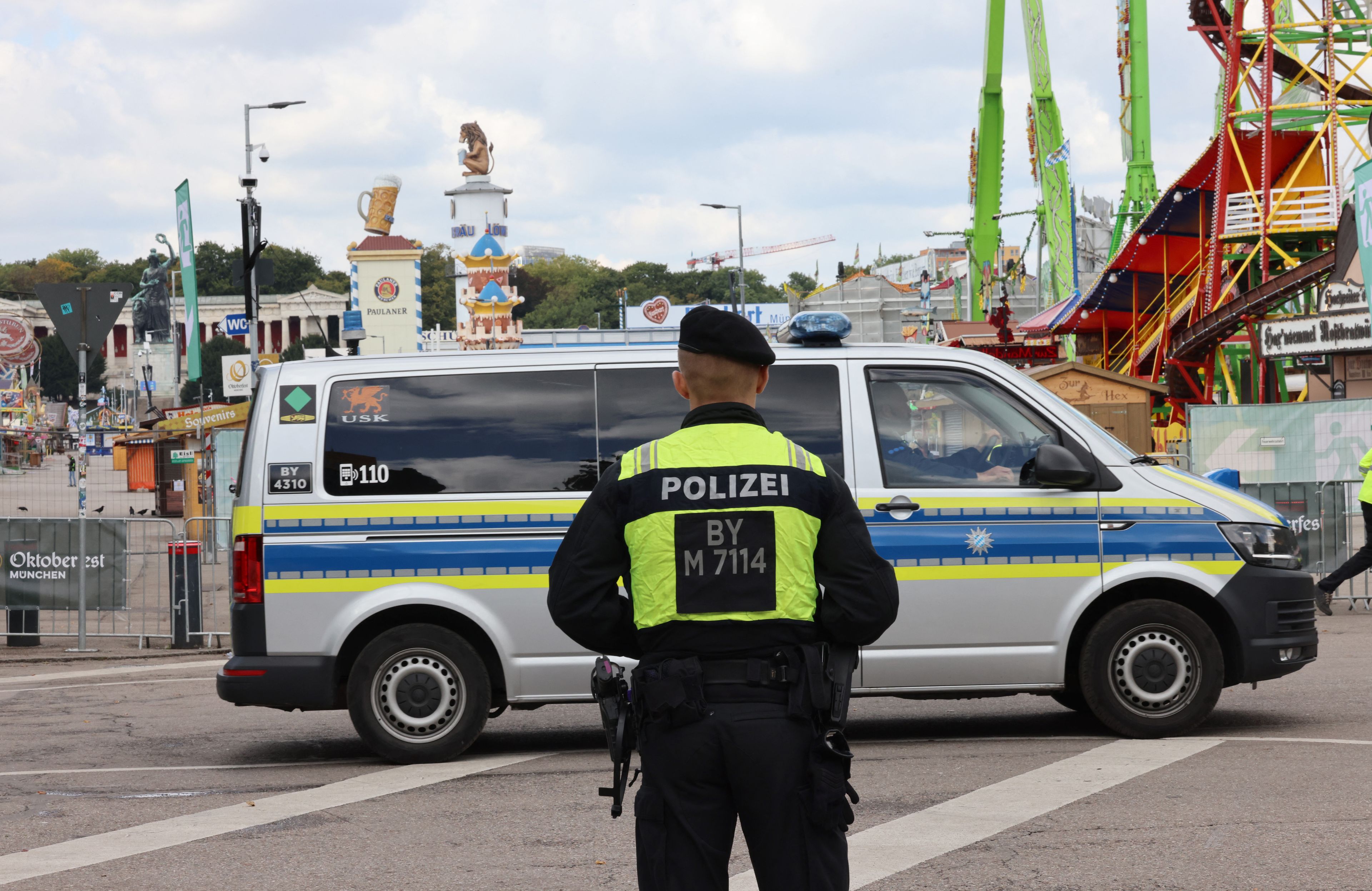 Policjant w kamizelce z napisem „Polizei” stoi przed radiowozem na tle wesołego miasteczka i dekoracji piwnych podczas Oktoberfest w Monachium. Festiwal pozostaje zamknięty w związku z alarmem bombowym