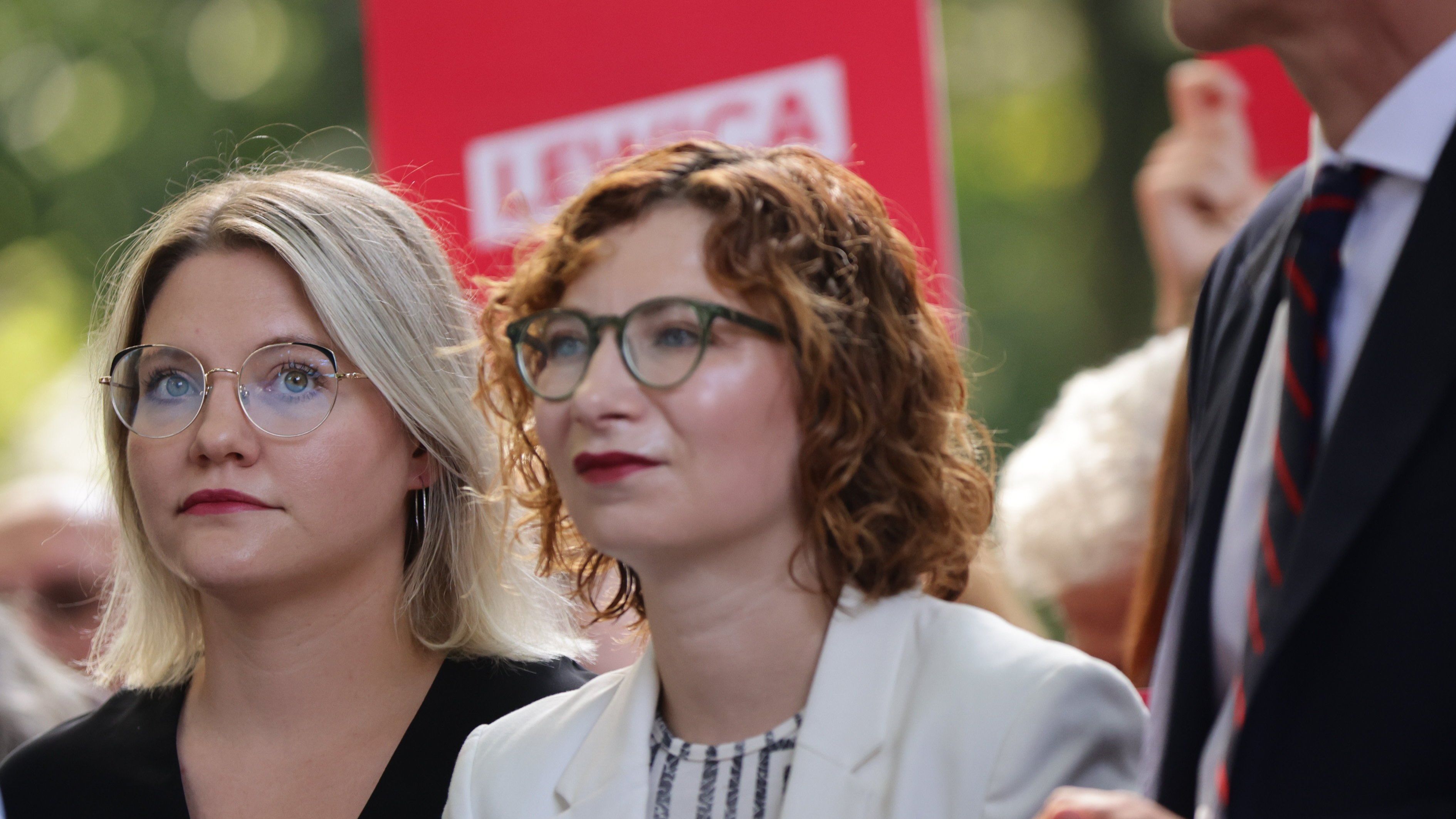 Dwie elegancko ubrane kobiety patrzą przed siebie. Za nimi baner z napisem: Lewica