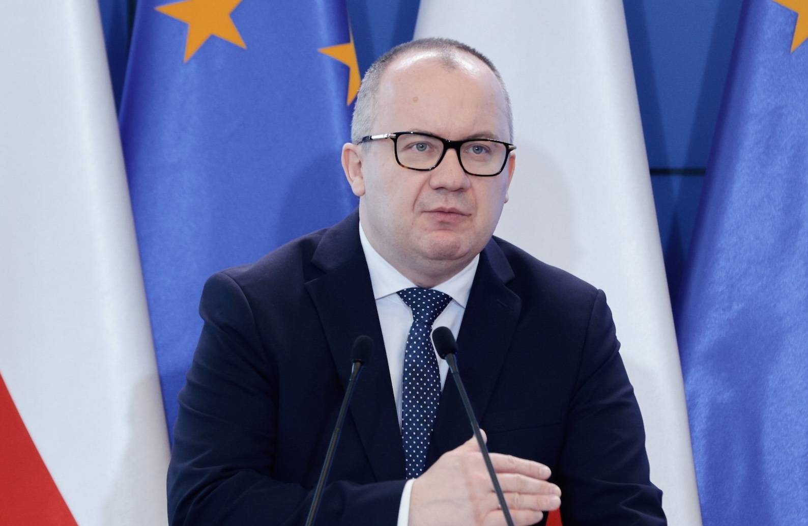 Minister Adam Bodnar przemawia do mikrofonu na tle polskich i unijnych flag. Ma na sobie ciemny garnitur i granatowy krawat w kropki.