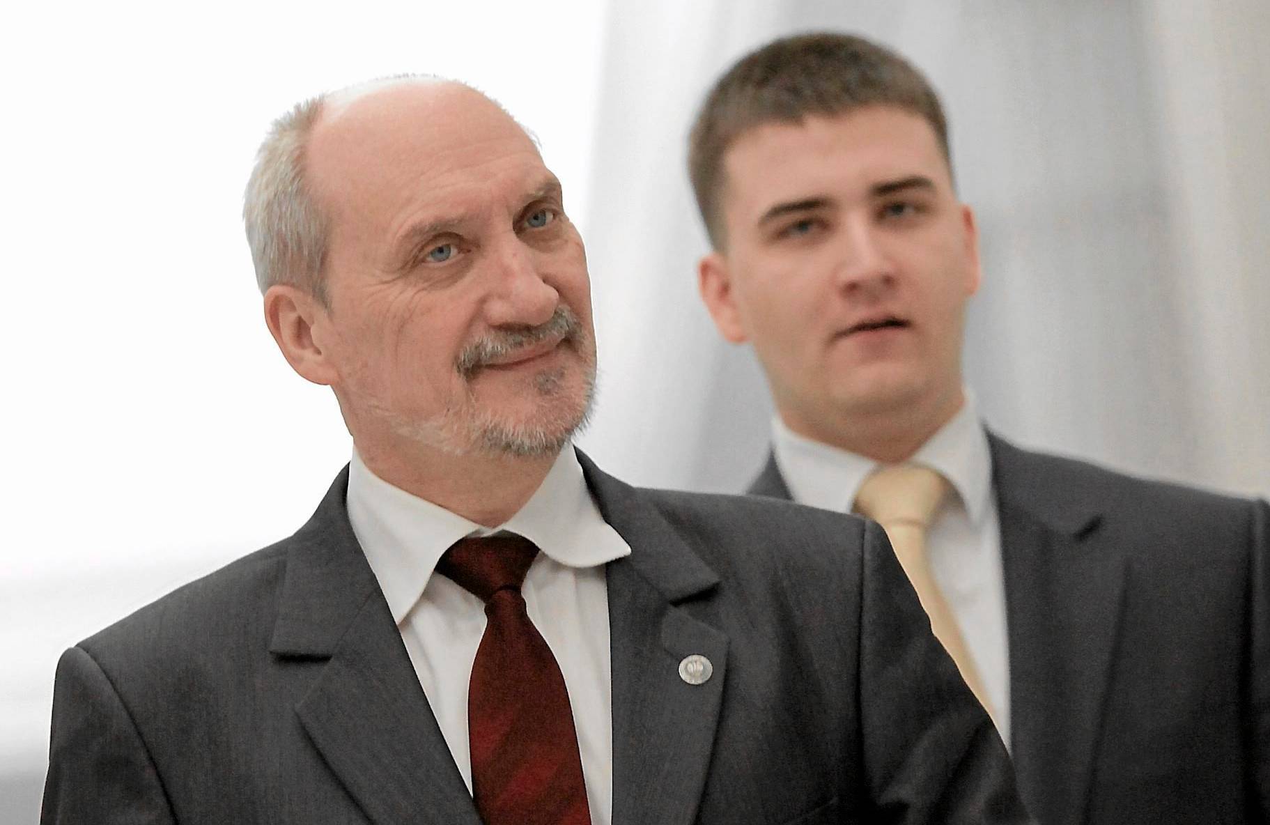 Łysiejący mężczyzna z brodą, w garniturze i białej koszuli, za nim młody mężczyzna w garniturze i białej koszuli