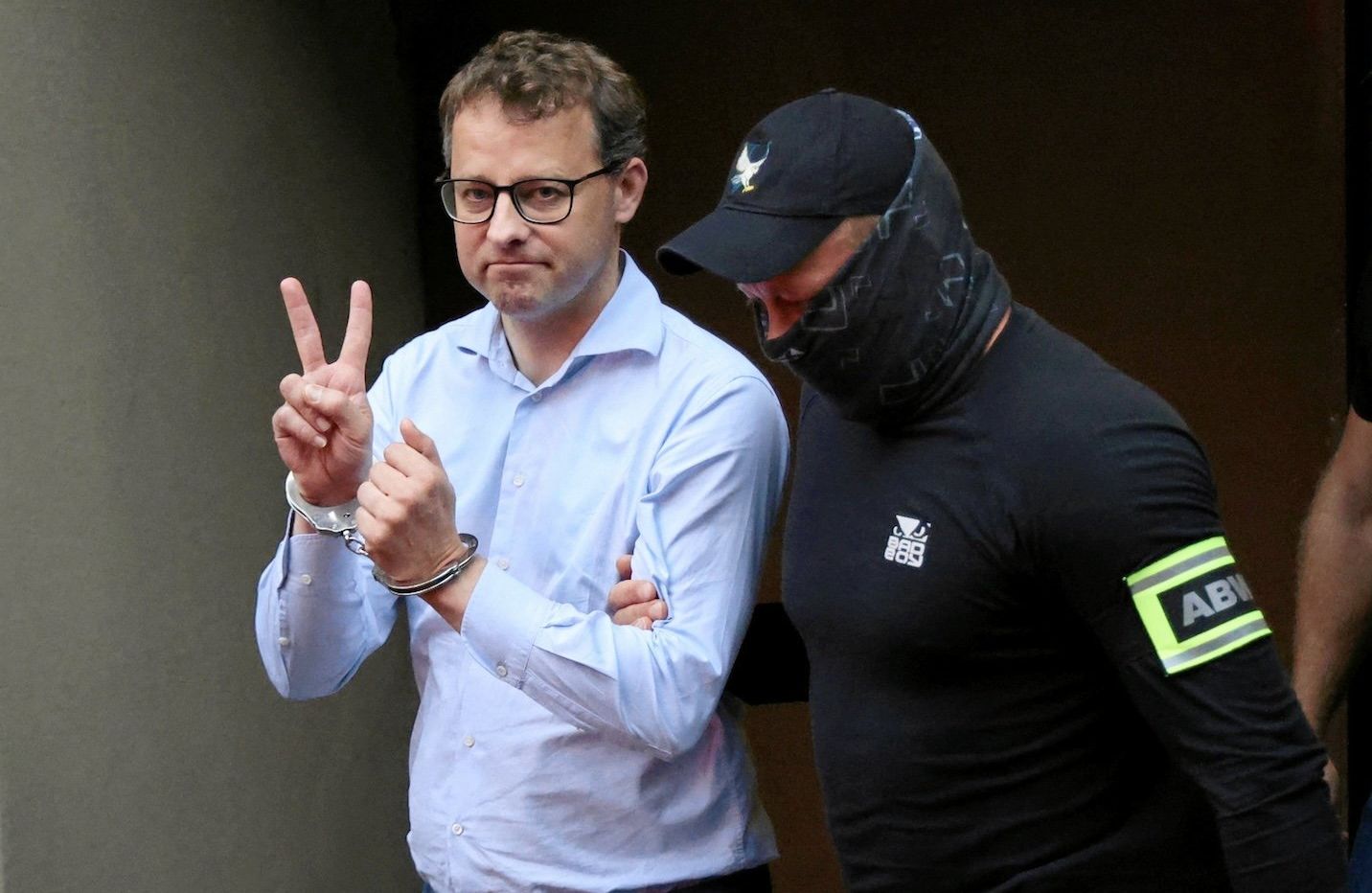 Marcin Romanowski wyprowadzany w kajdankach przez funkcjonariusza ABW. Unosi rękę w geście V.