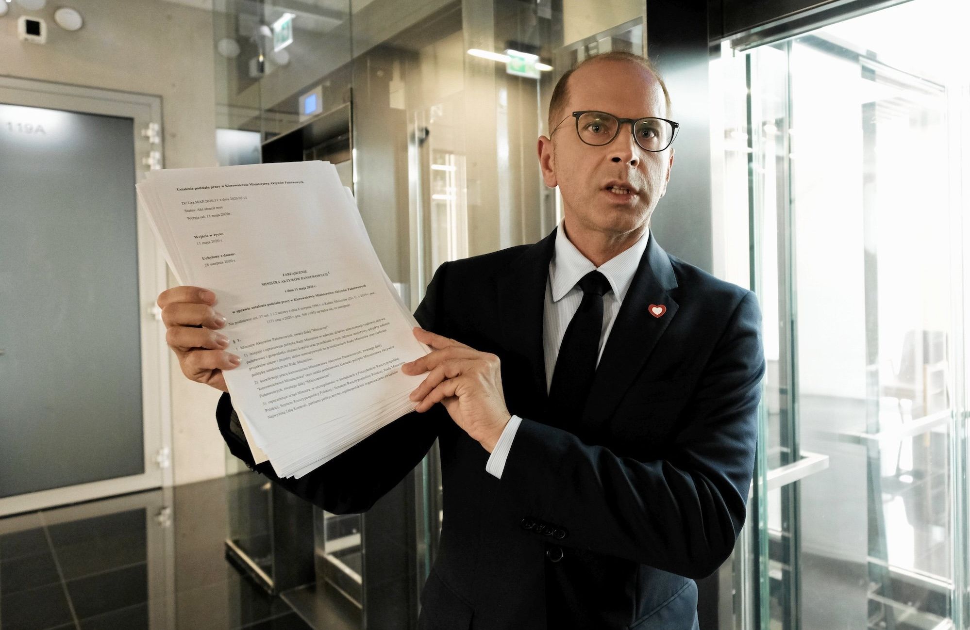 Michał Szczerba w budynku Sejmu pokazuje plik dokumentów. Ubrany jest w ciemny garnitur i krawat