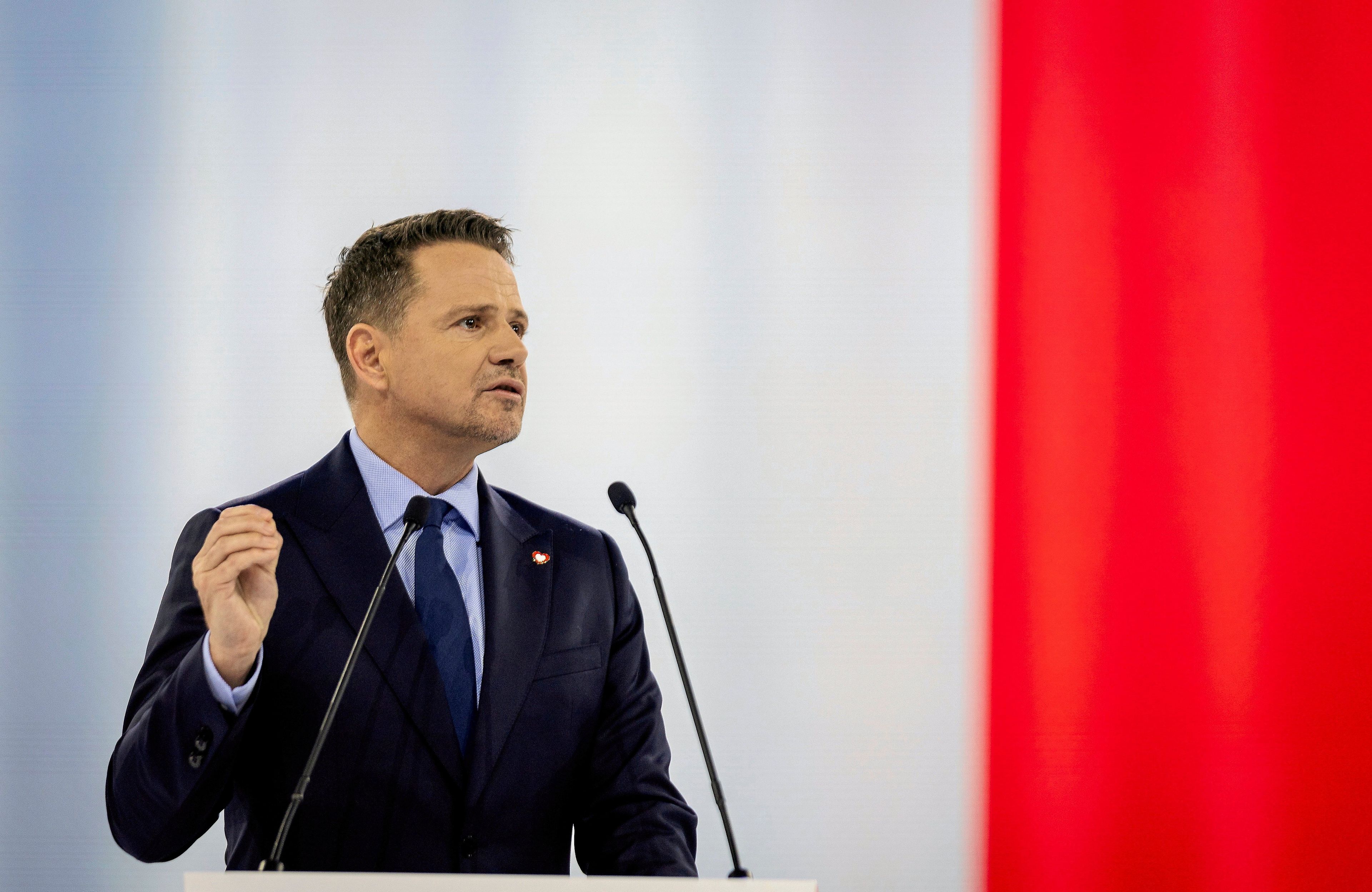 Rafał Trzaskowski przemawia zza mównicy na tle biało-czerwonej flagi