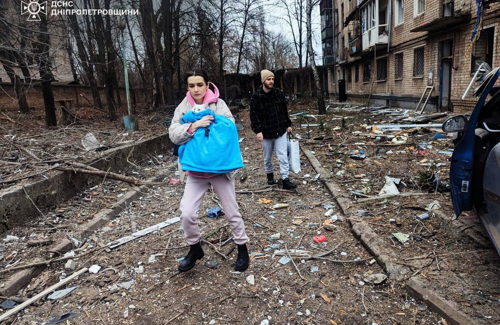 Na zdjęciu są dwie osoby: kobieta i mężczyzna. Są wśród ulicy, na ziemi widać fragmenty zniszczonych budynków. Kobieta trzyma w rękach białego kota, który jest owinięty w błękitny koc