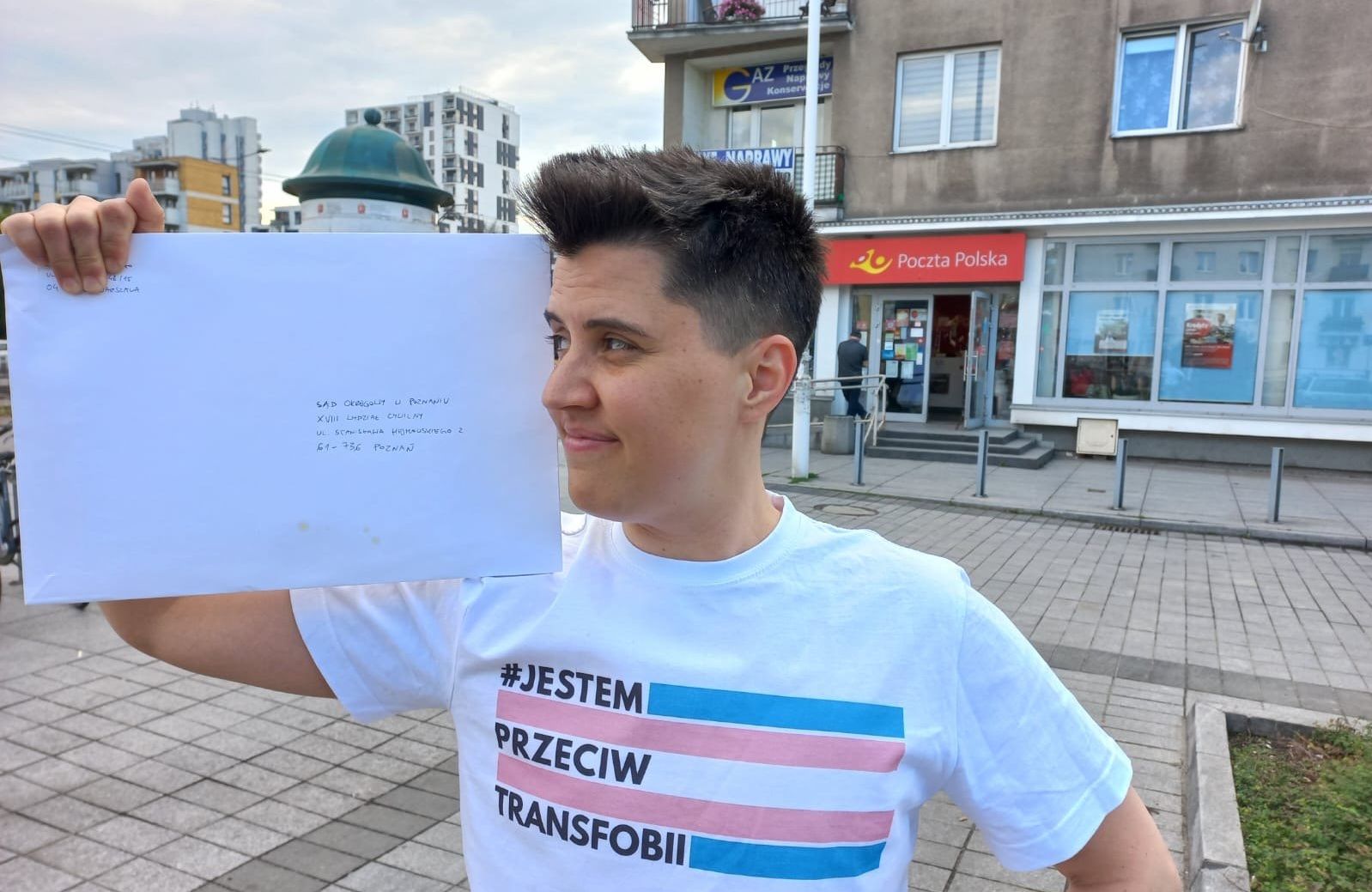 Młody mężczyzna z krótkimi ciemnymi włosami, ubrany w białą koszulkę z różowymi i błękitnymi paskami oraz napisem #JestemPrzeciwTransfobii pozuje z białą kopertą formatu A4. Za nim napis "Poczta Polska" na czerwonym tle.