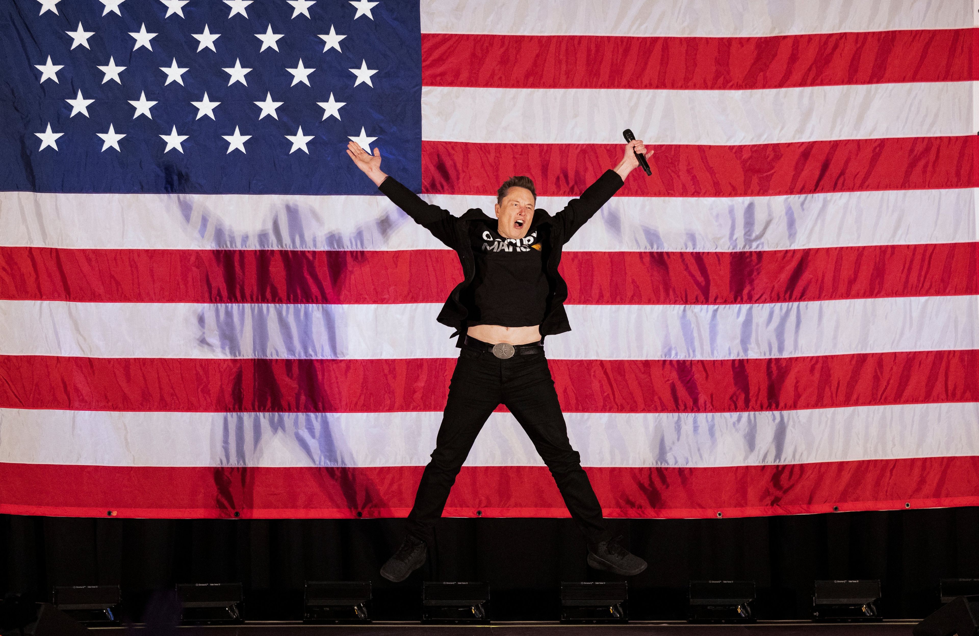 18.10.2024, Oaks, Pensylwania, USA. Elon Musk skacze na scenie na tle wielkiej amerykańskiej flagi podczas wiecu zwolenników Donalda Trumpa zorganizowanego przez założoną przez niego organizację America PAC
