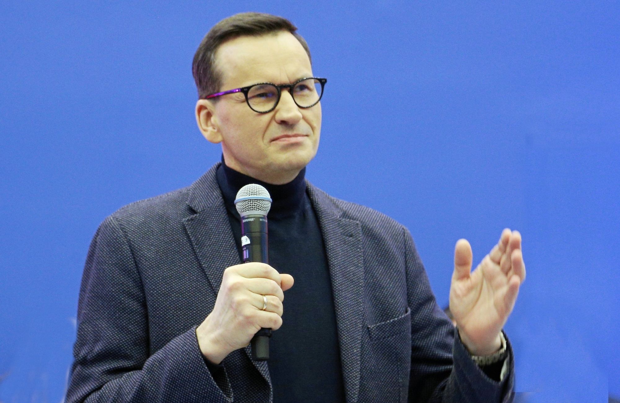 Mateusz Morawiecki przemawia, w ręku trzyma mikrofon. Ubrany jest w czarny golf i szarą marynarkę.