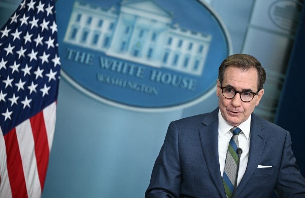 mężczyzna w okularach i garniturze na tle logo Białego Domu i flagi USA