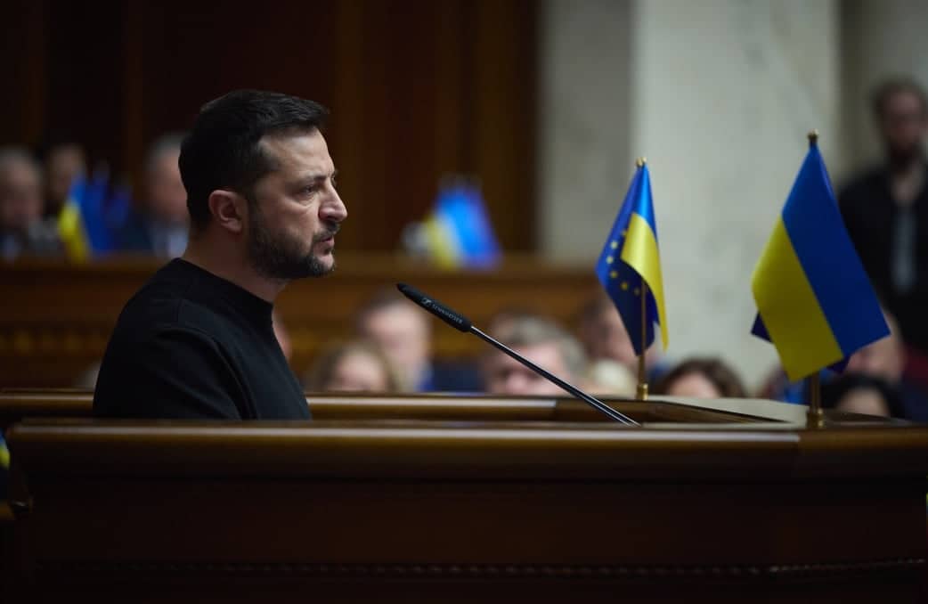 Na zdjęciu jest mężczyzna ok. 45 lat. Ma czarne krótkie włosy i brodę. Jest ubrany w czarną bluzę. Stoi przy ambonie. Po prawej stronie na ambonie są małe flagi- ukraińska oraz Unii Europejskiej