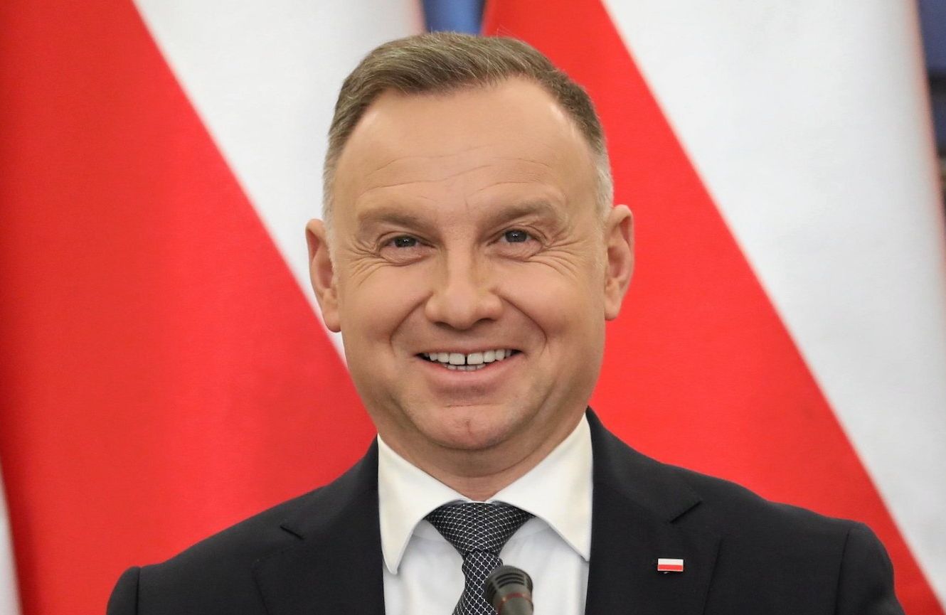 Szeroko uśmiechnięty prezydent Andrzej Duda na tle biało-czerwonych flag. Na pierwszym planie bukiet z białych i czerwonych róż.