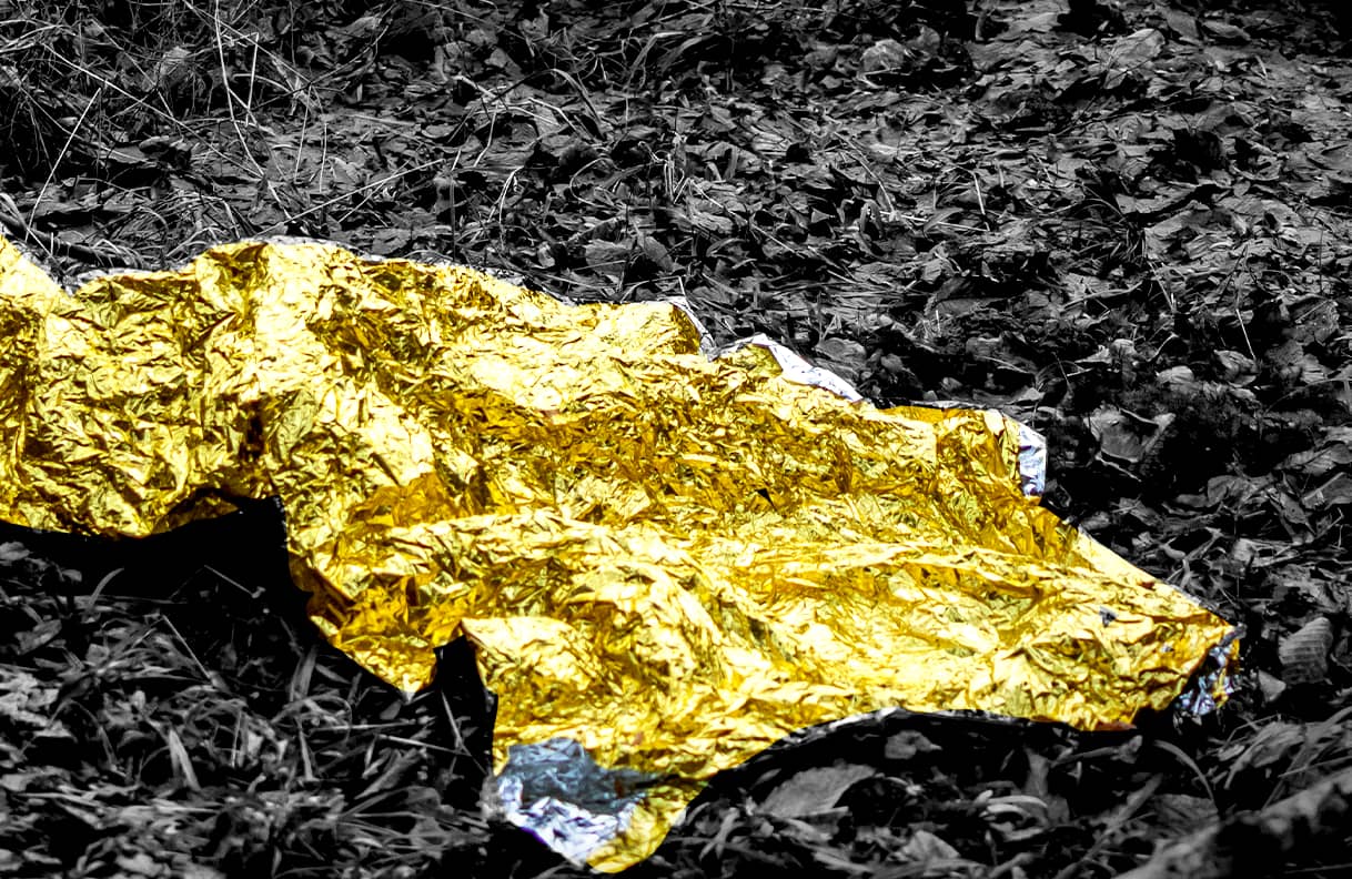 Na pierwszym planie pognieciony złoty koc termiczny, w tle fragment lasu.