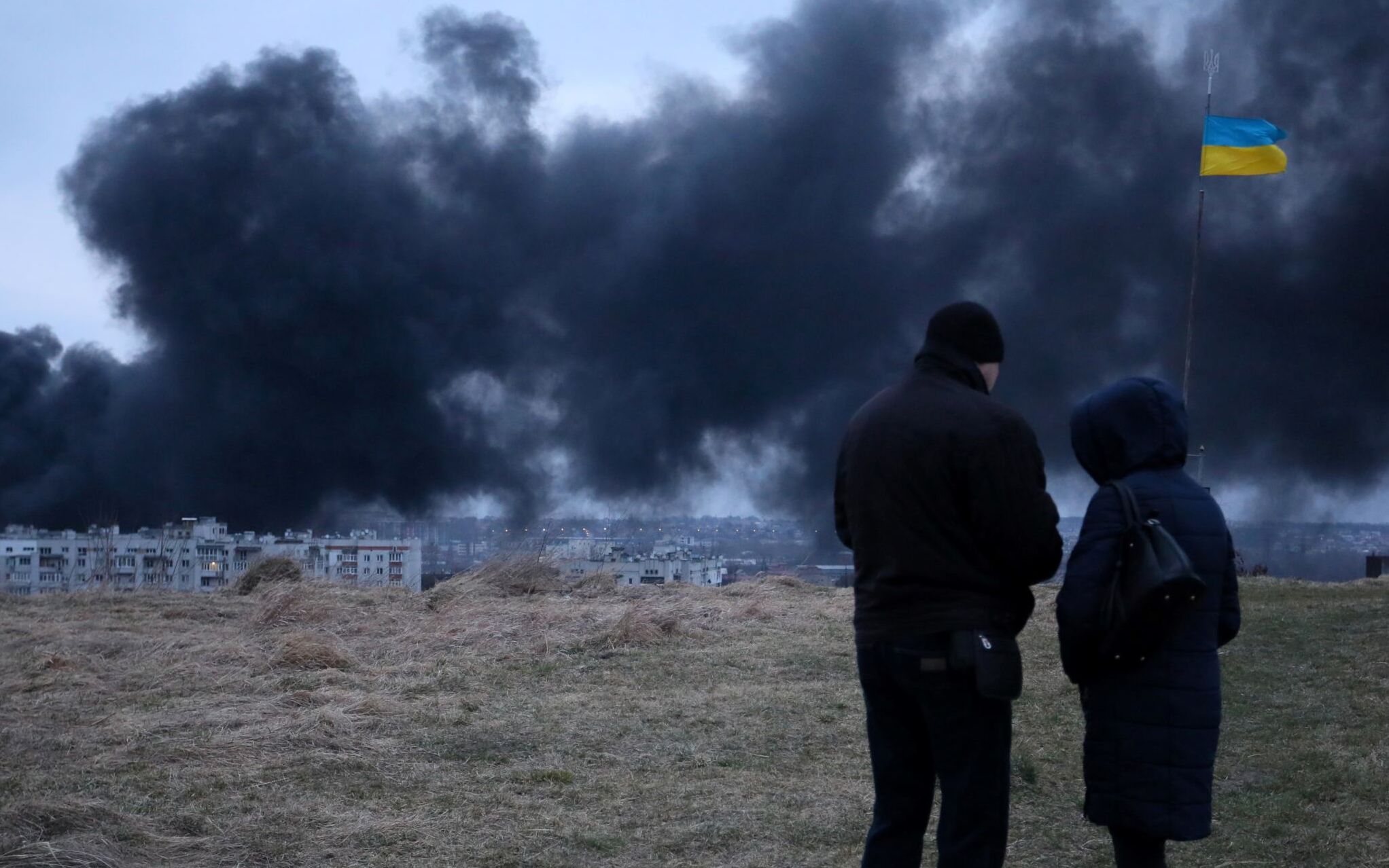 People stand in front of an Ukrainian national flag fluttering as dark smoke and flames rise from a fire following an air strike in the western Ukrainian city of Lviv, on March 26, 2022. - At least five people wounded in two strikes on Lviv, the regional governor said, in a rare attack on a city that has escaped serious fighting since Russian troops invaded last month. (Photo by Aleksey Filippov / AFP)