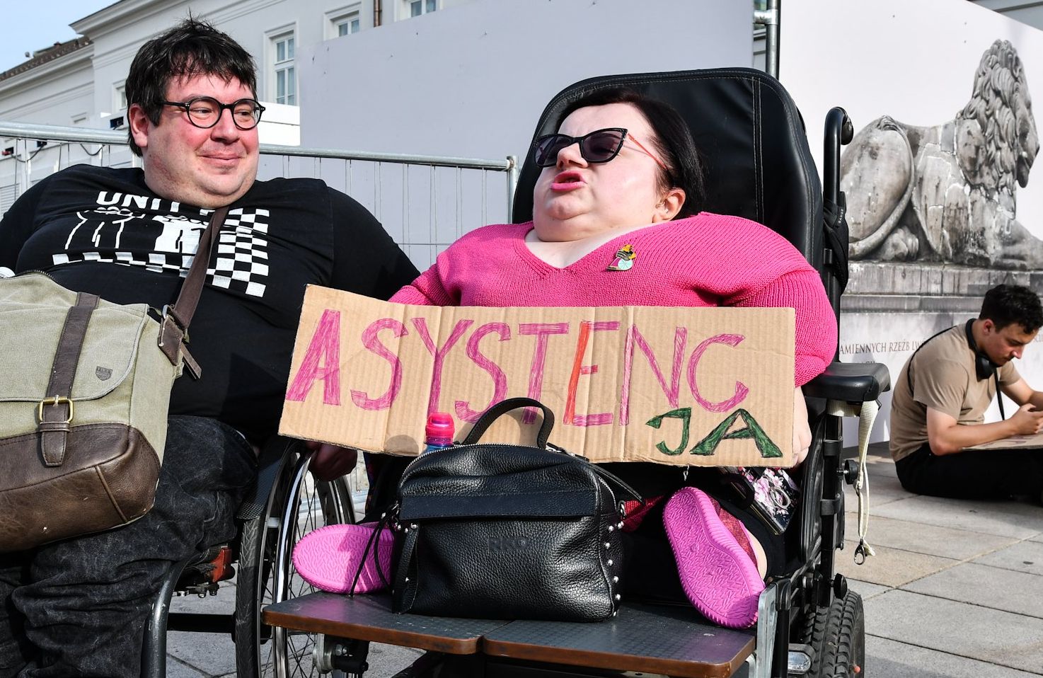 Dwie osoby z niepełnosprawnością na wózkach przed pałacem prezydenckim. Jedna trzyma transparent z napisem "asystenc-ja"