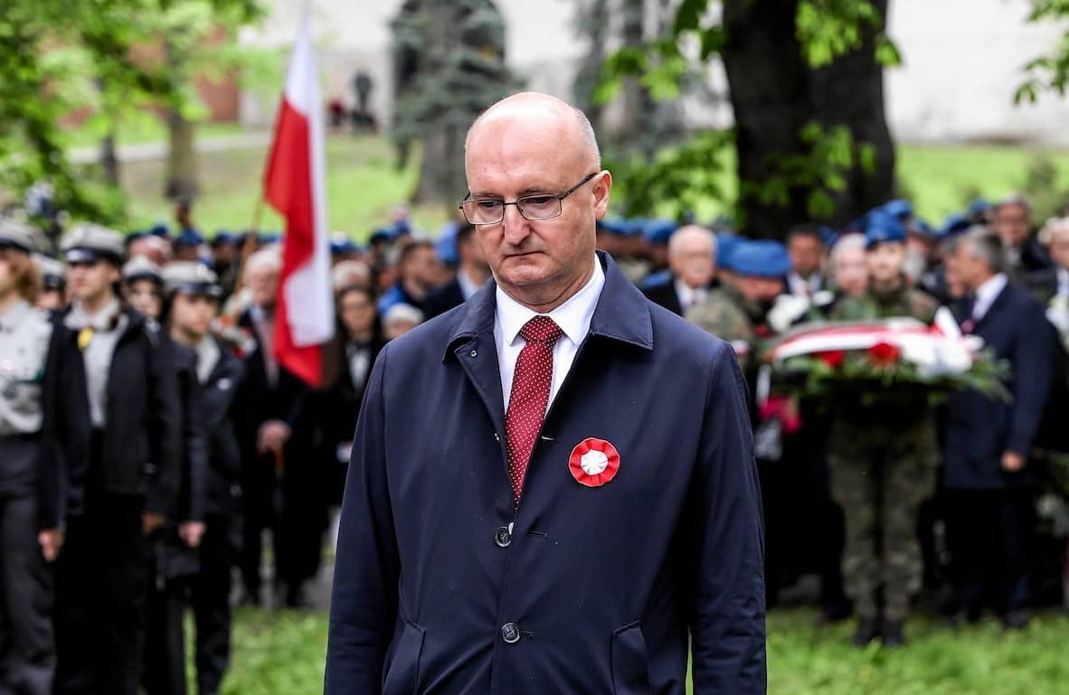 Wiceminister Piotr Wawrzyk na obchodach święta Konstytucji 3 Maja w Kielcach. Stoi ubrany w ciemny garnitur i płaszcz i spogląda ponuro w dół. W tle zgromadzenie ludzi z biało-czerwonymi flagami.