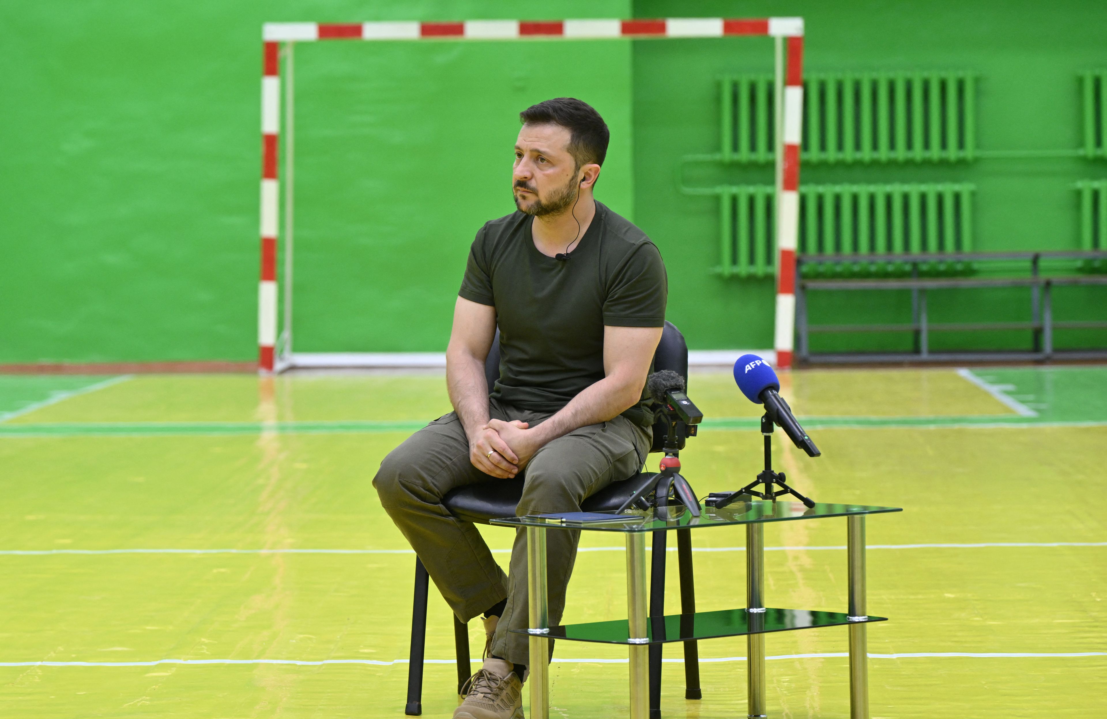 Mężczyzna w ciemnozielonej koszulce siedzi na krześle na środku sali gimnastycznej. Przed nim na małym stole mikrofony mediów