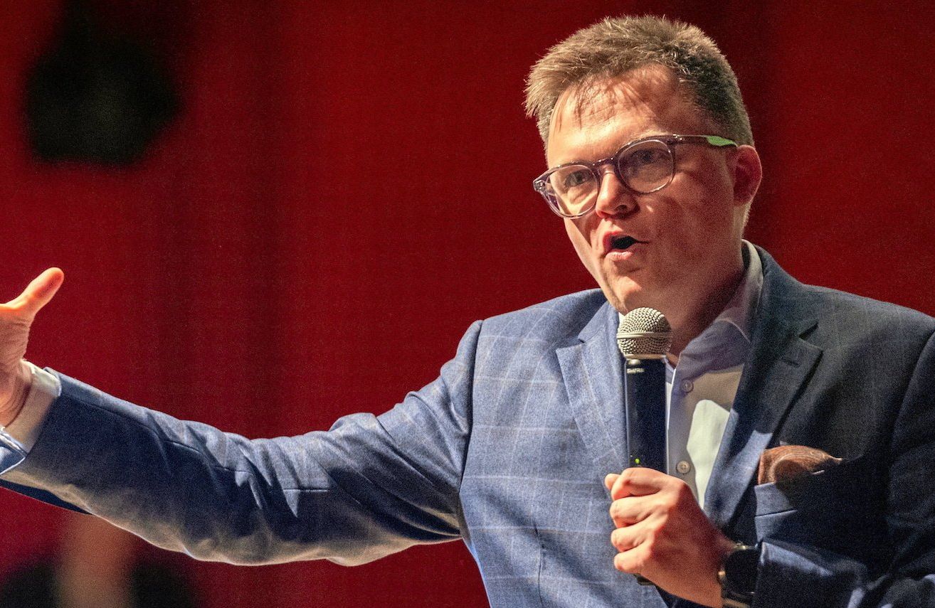 Szymon Hołownia podczas przemówienia. Ubrany w garnitur w kratkę unosi prawą rękę.
