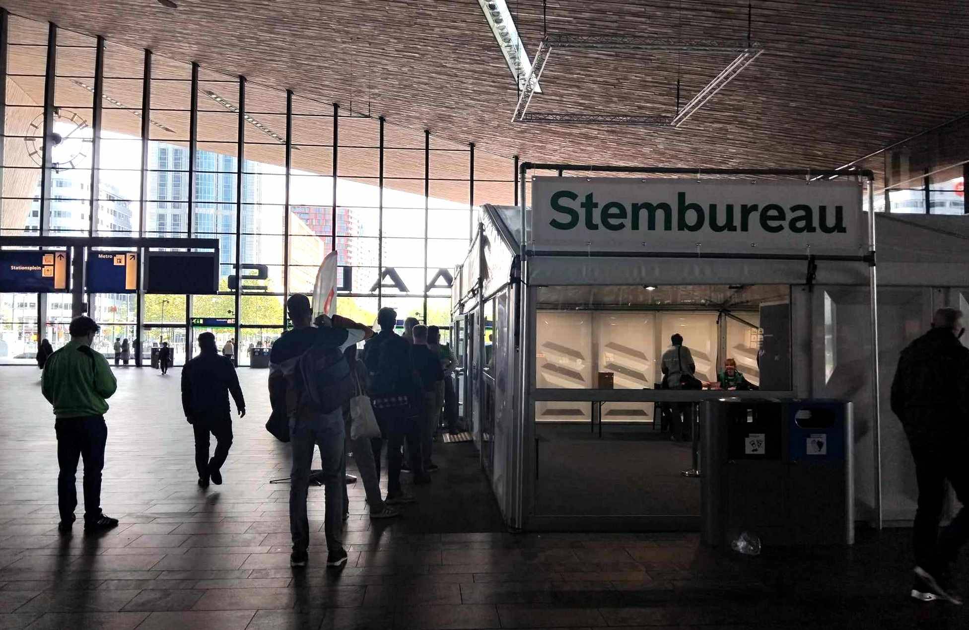 Przestronna hala główna dworca kolejowego w Rotterdamie - brązowe, asymetryczne sklepienie i przeszklona ściana z wejściem, na tle tej ściany po prawej stronie ustawiony tymczasowy lokal wyborczy; przy lokalu nieduża kolejka osób, które czekają, żeby oddać głos w wyborach w Holandii