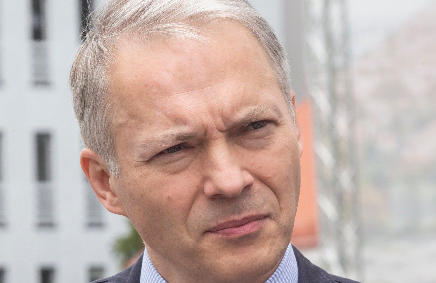 Jacek Żalek, mężczyzna z siwymi włosami w szarym garniturze i niebieskiej koszuli, w klapie marynarki ma przypinkę w kształcie czerwonej róży. Wiceminister spogląda w lewo i mruży oczy pod światło.