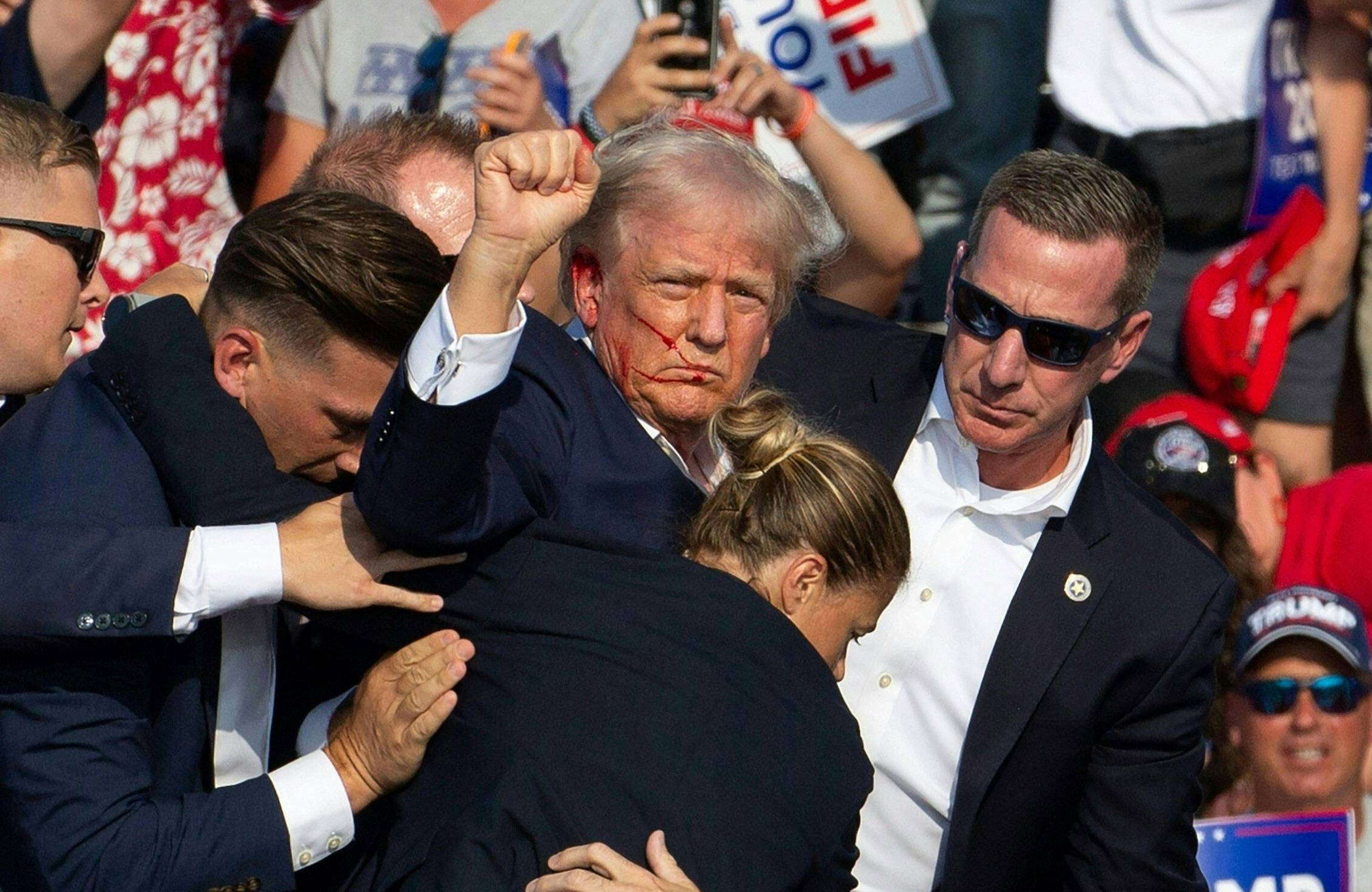 Donald Trump w otoczeniu ochroniarzy, z zakrwawioną twarzą, podnosi pięść.