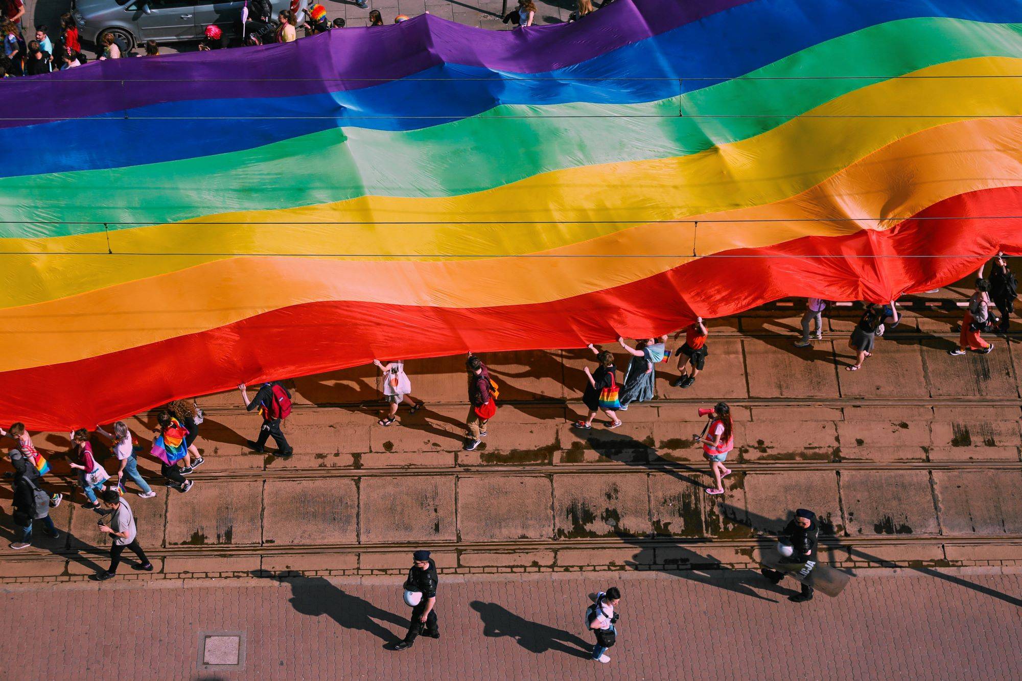 Manifestanci idą ulicą niosąc wielką tęczową flagę