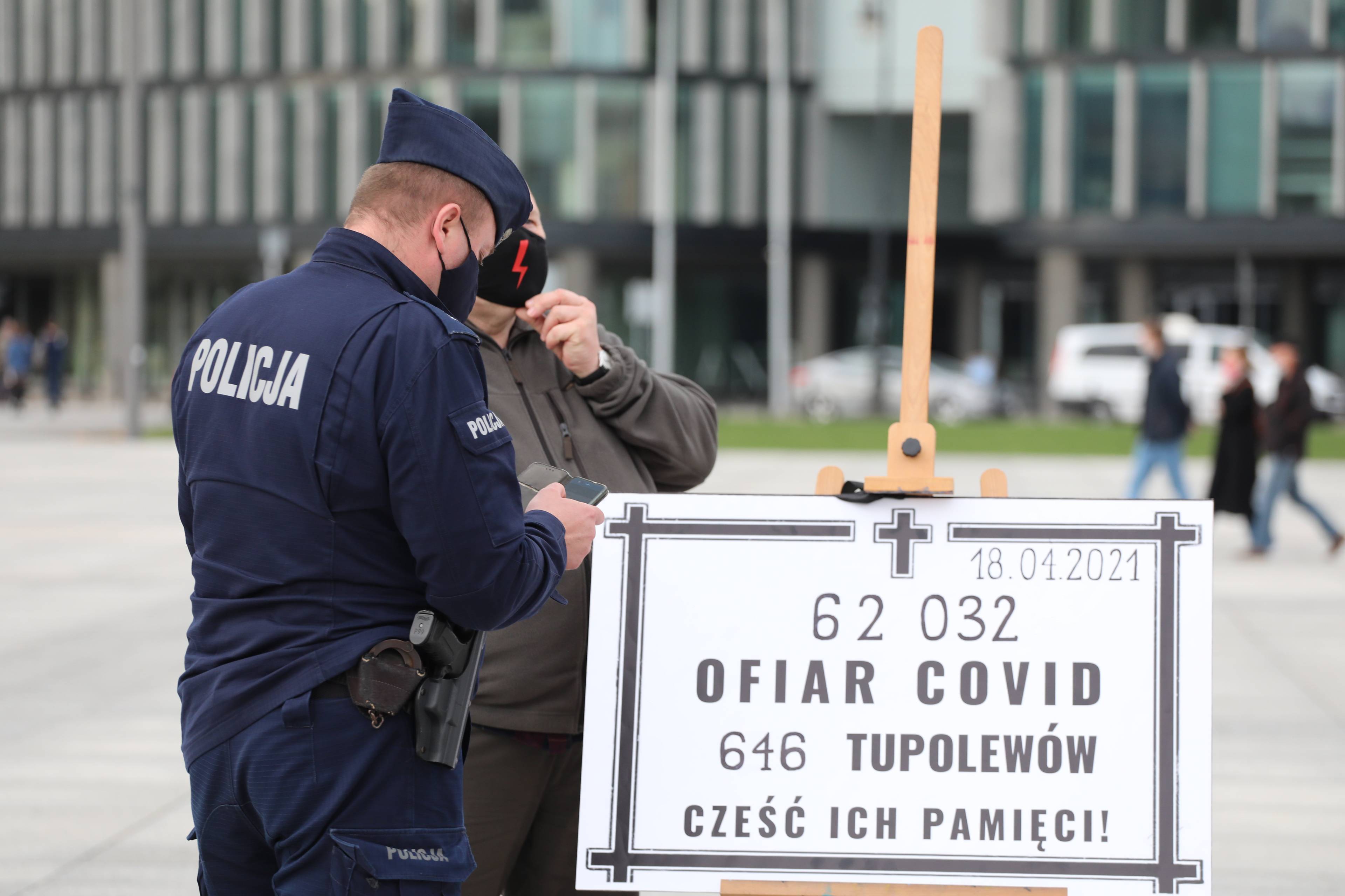 Policjant stoi przed tablicą ustawioną na Placu Piłsudskiego z napisem „62 032 ofiar Covid 616 tupolewów Cześć Ich Pamięci”