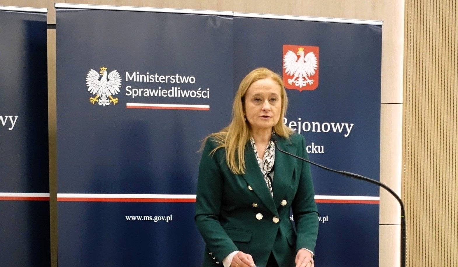 Kobieta, długie blond wóly porzemawia na tle logo ministerstwa sprawiedliwości