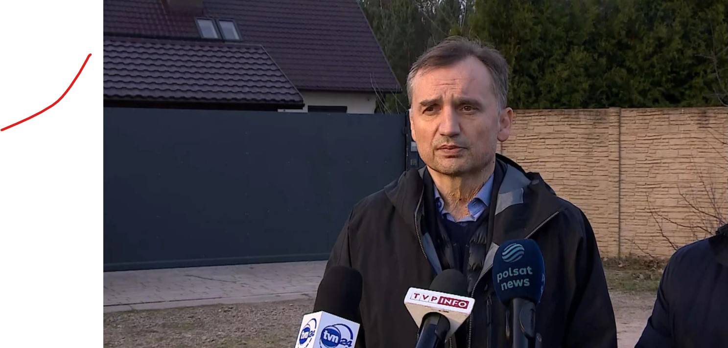 Mężczyzna z krótkimi ciemnymi włosami stoi przed ogrodzonym wysokim płotem domem i rozmawia z dziennikarzami