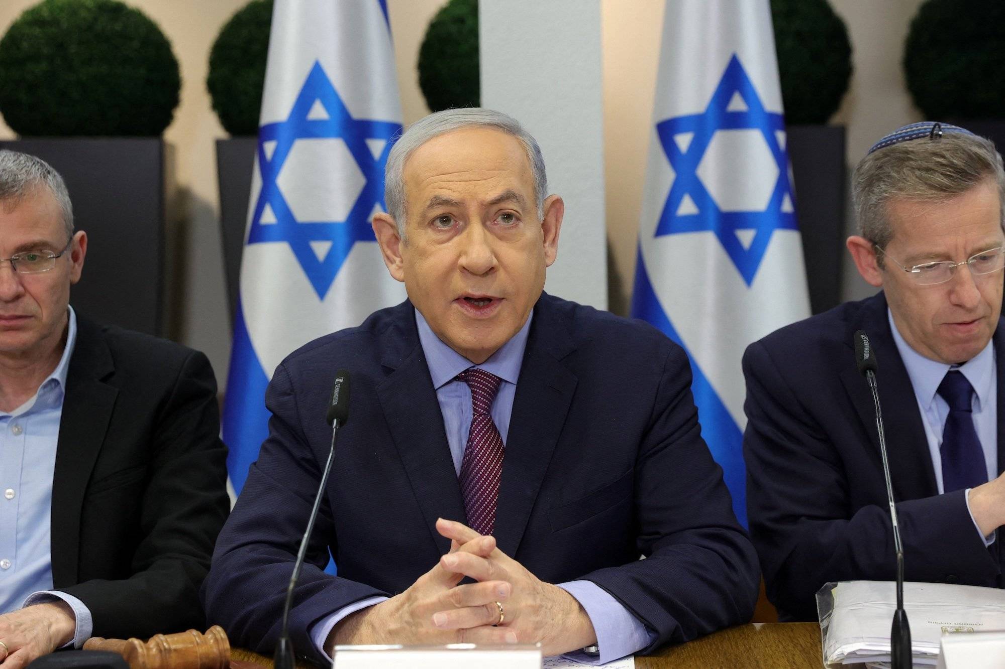 Binjamin Netanjahu przemawia na konferencji na tle flag Izraela. Siedzi za stołem i opiera na nim przedramiona, ma splecione palce dłoni.