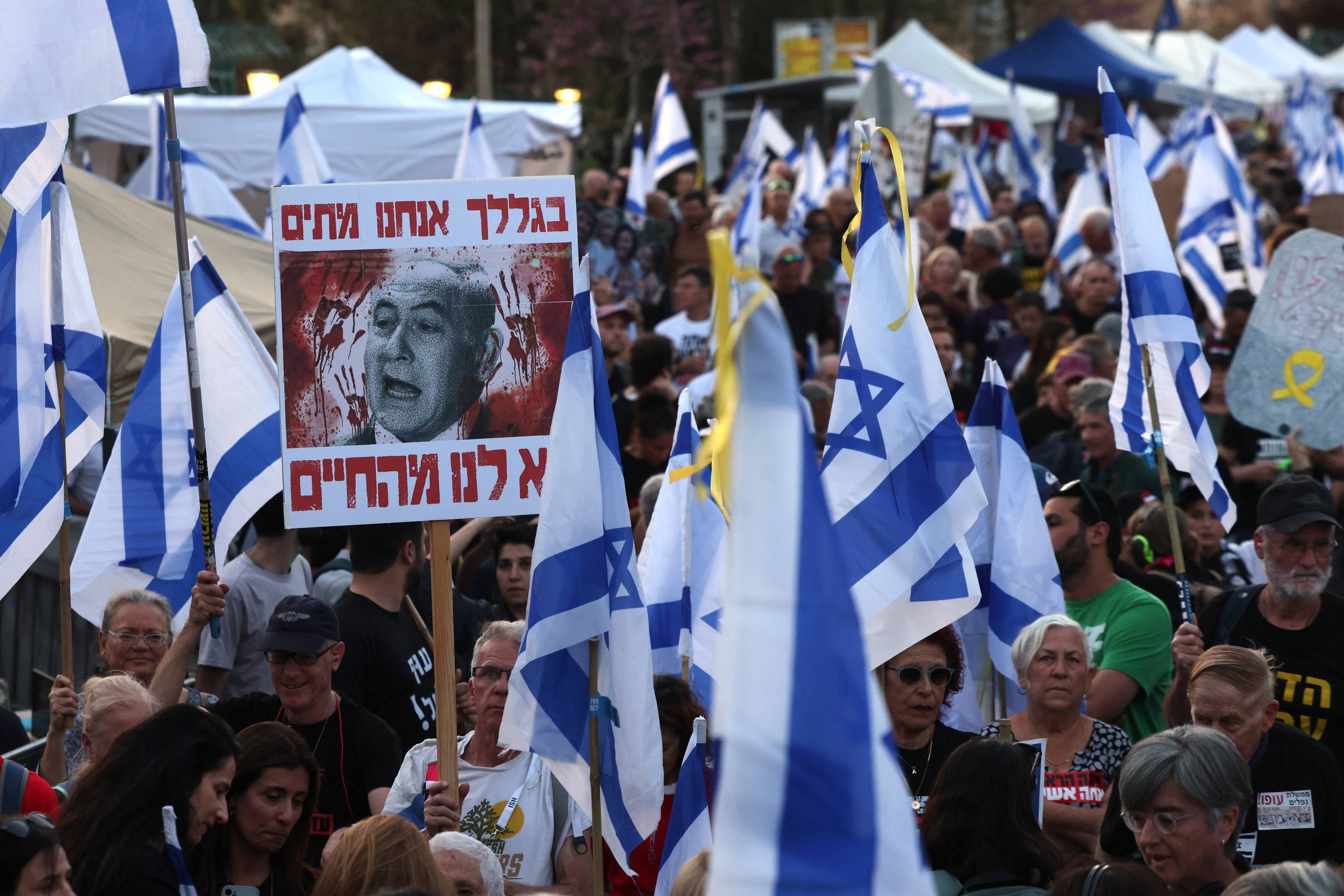 Protest w Izraelu, nad tłumem izraelskie flagi i portret Beniamina Netanjahu z domalowanymi śladami krwi