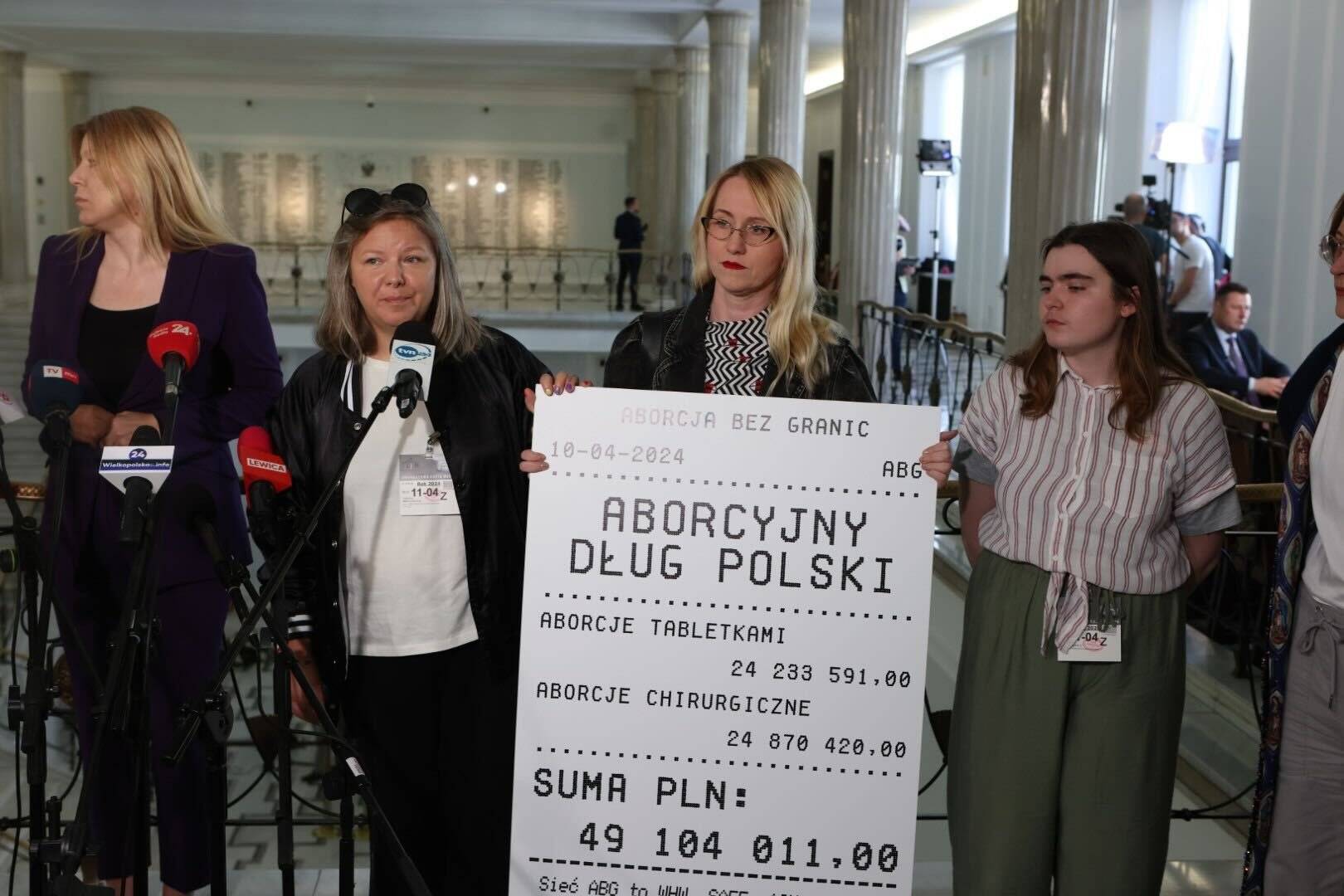 Działaczki Aborcji bez Granic stoją w holu Sejmu podczas konferencji prasowej. Jedna z nich trzyma powiększony paragon z napisem aborcyjny dług Polski
