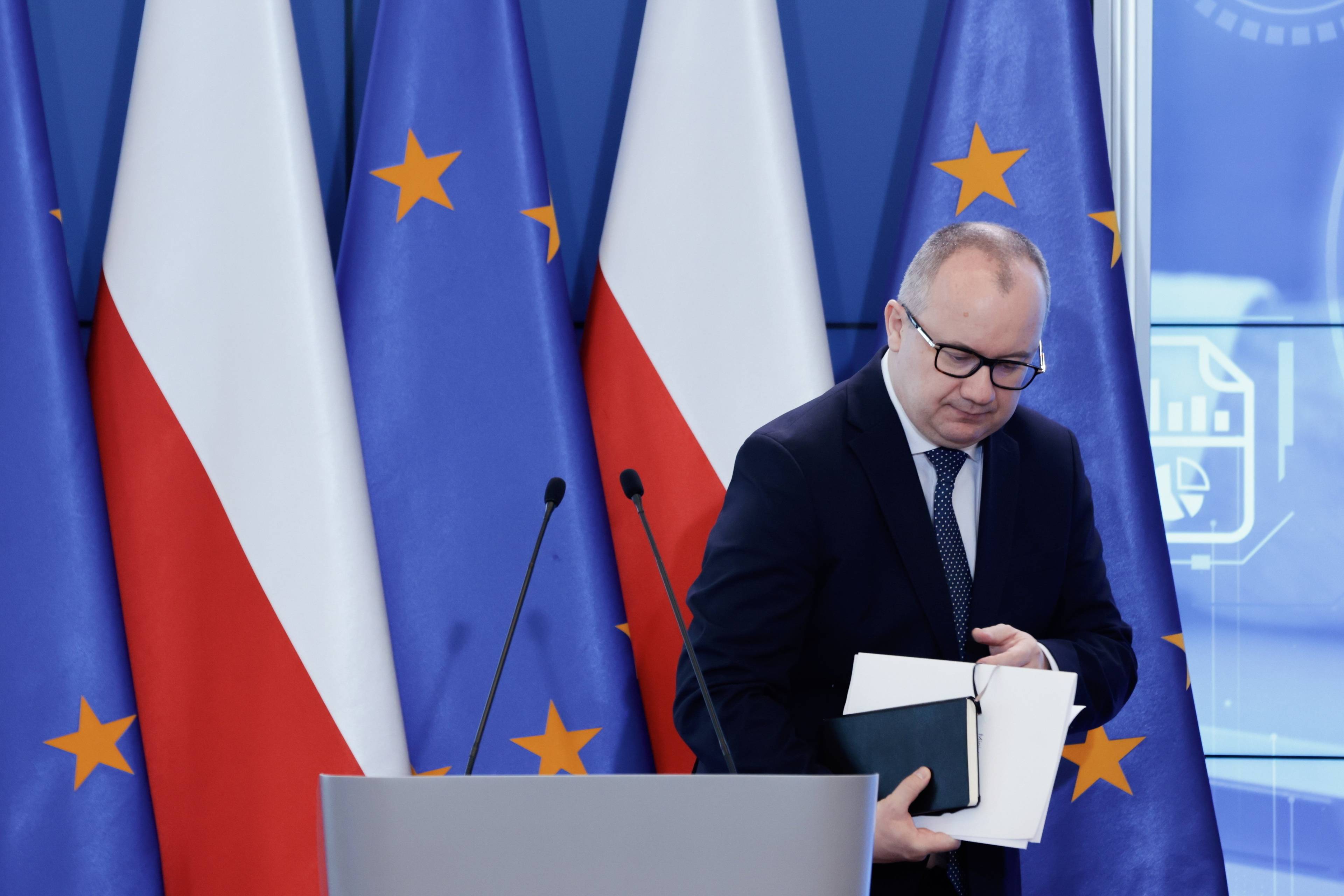 Adam Bodnar schodzi z mównicy, w ręce trzyma notes i papiery, za nim flagi Polski i UE