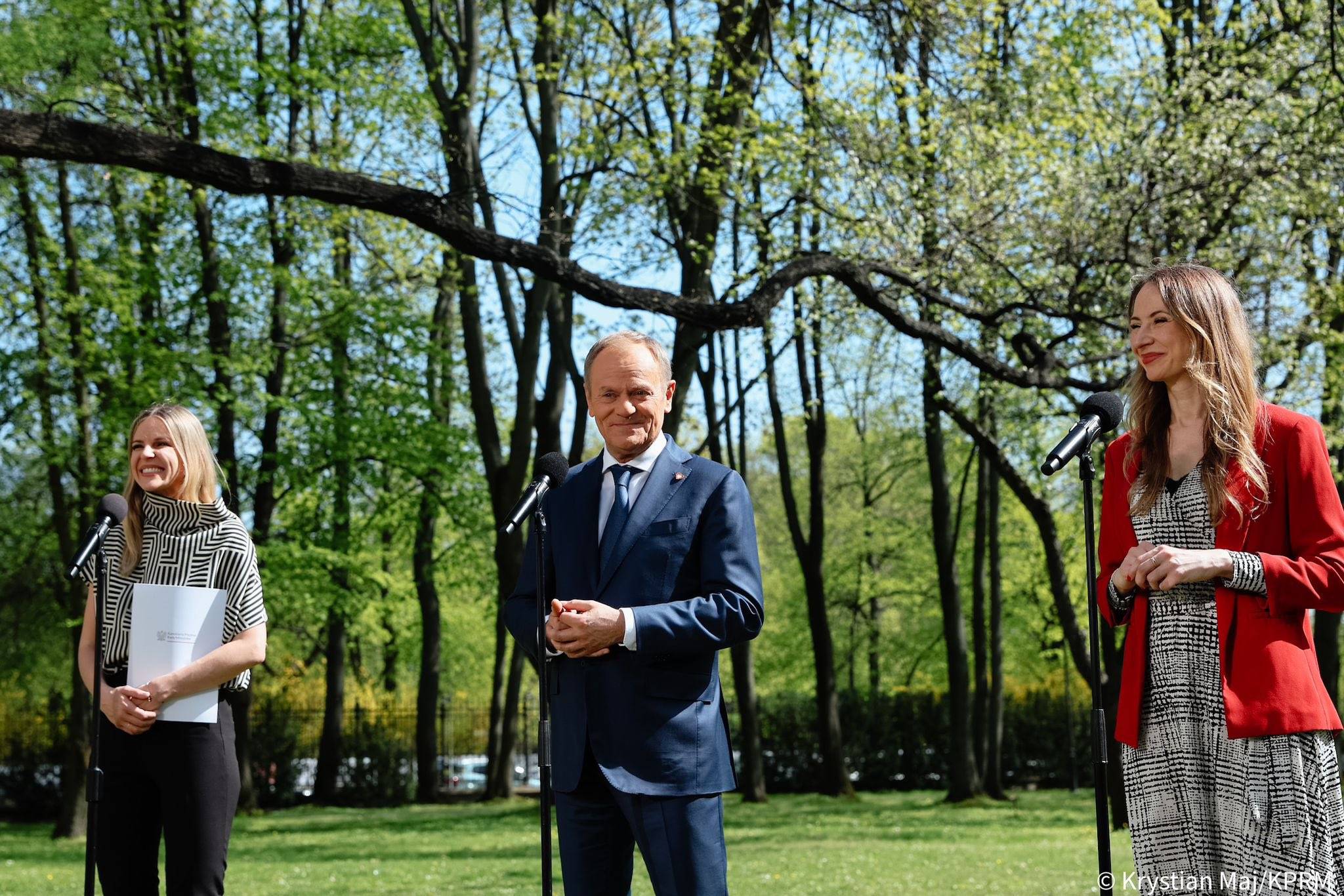 na zdjęciu widzimy stojących przed mikrofonami w zielonym słonecznym parku - premiera Donalda Tuska, ministrę pracy Agnieszkę Dziemianowicz-Bąk i sekretarz stanu Aleksandrę Gajewską