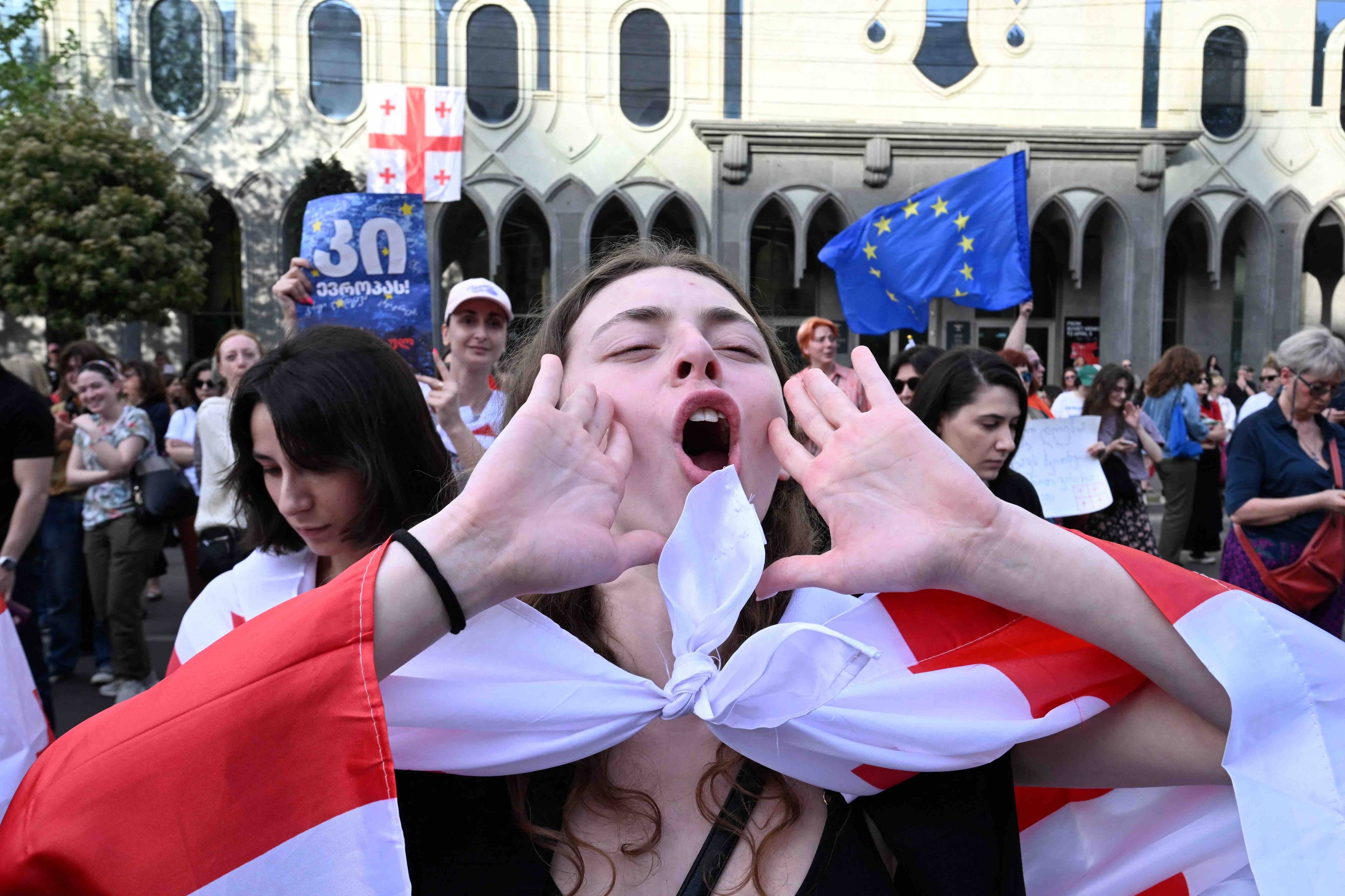 Uczestniczka demonstracji krzyczy, przykładając dłonie do ust, na ramionach ma gruzińską flagę narodową, w tle inni demonstranci z flagami gruzińskimi i UE. Gruzja