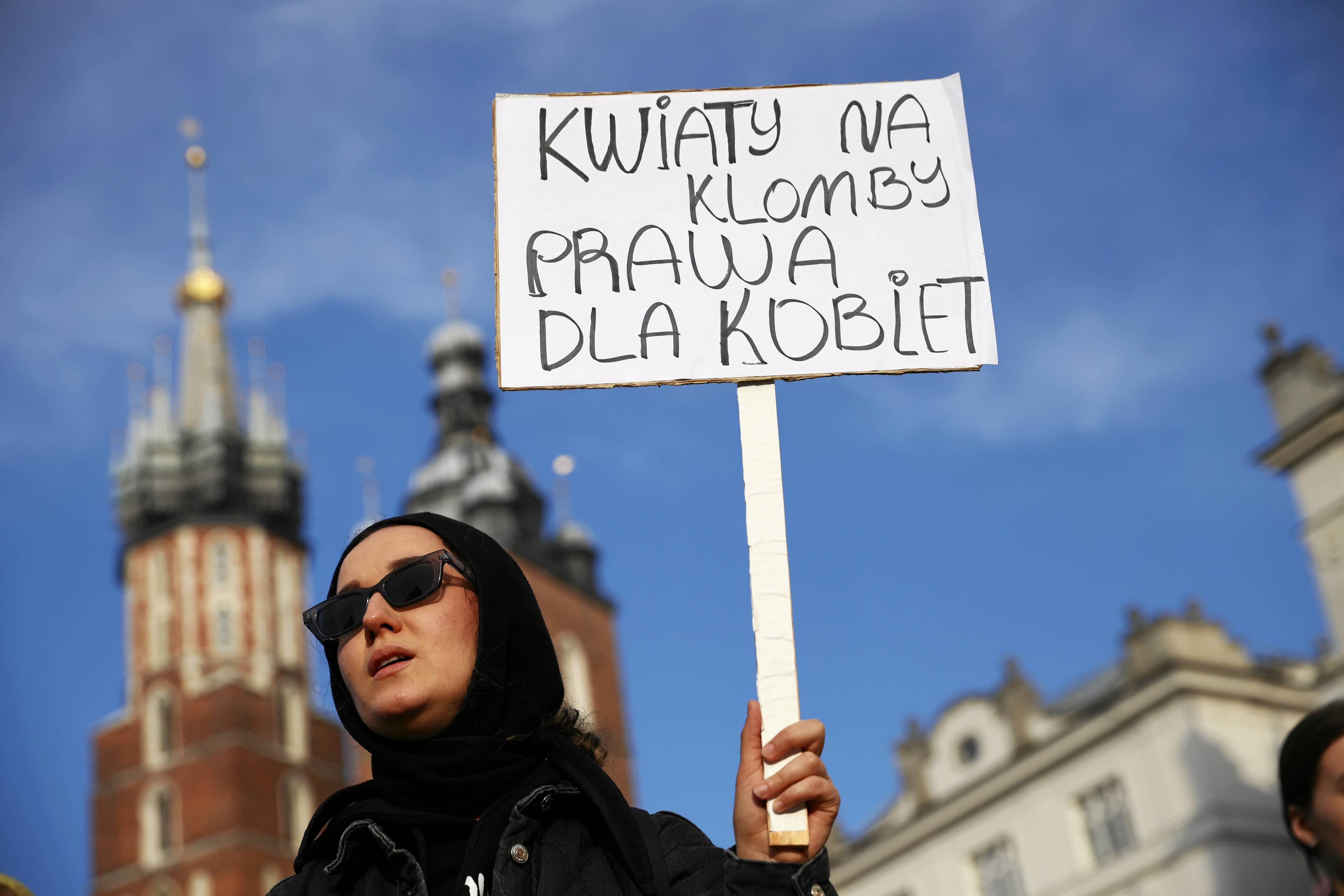 kobieta w ciemnych okularach i czarnej chustce trzyma transparent z napisem „Kwiaty na klomb, prawa dla kobiet". W tle dwie nierówne wieże Kościoła Mariackiego