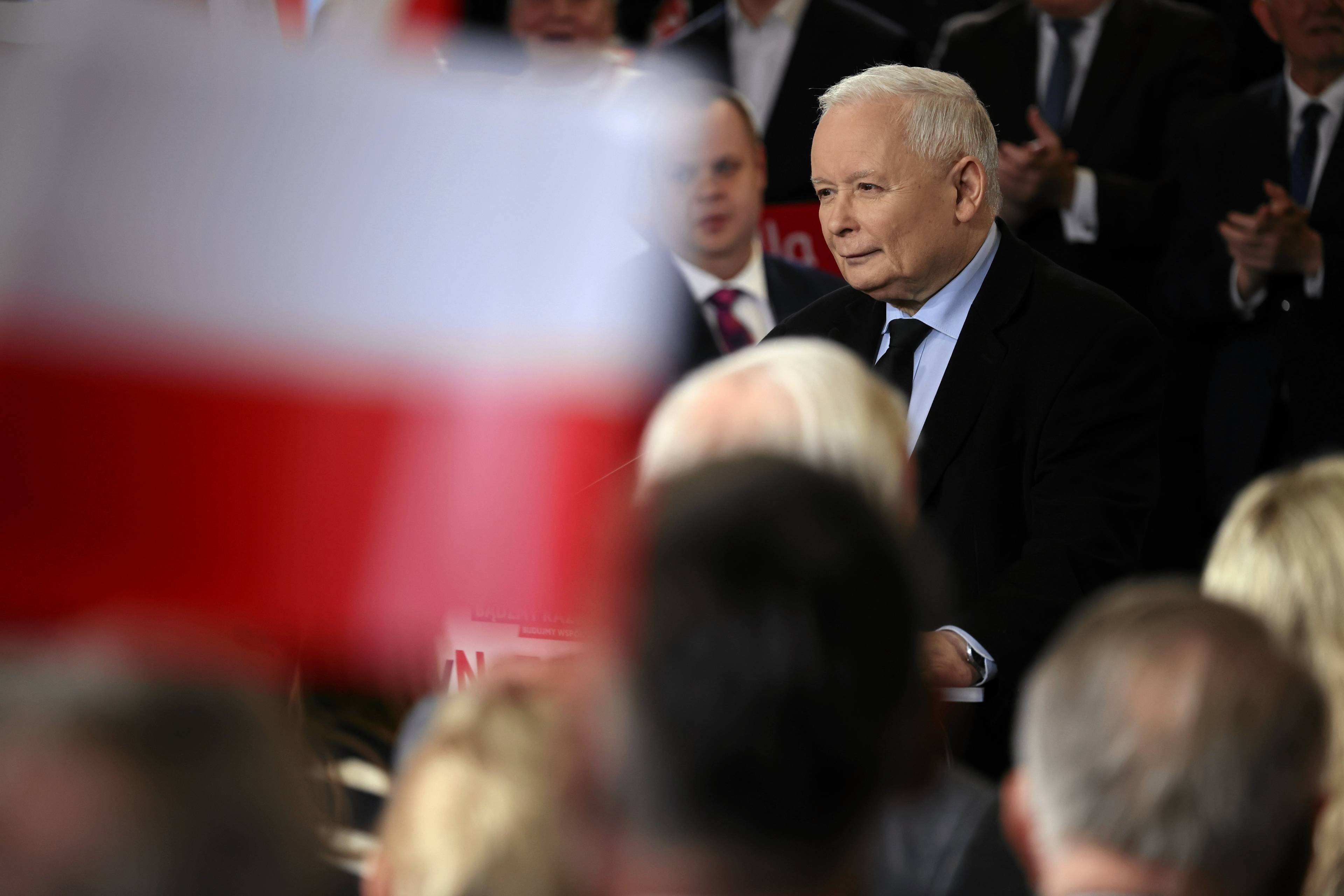 Jarosław Kaczyński w garniturze wśród ludzi na wiecu wyborczym, na pierwszym planie obok polityka lekko rozmyta flaga Polski