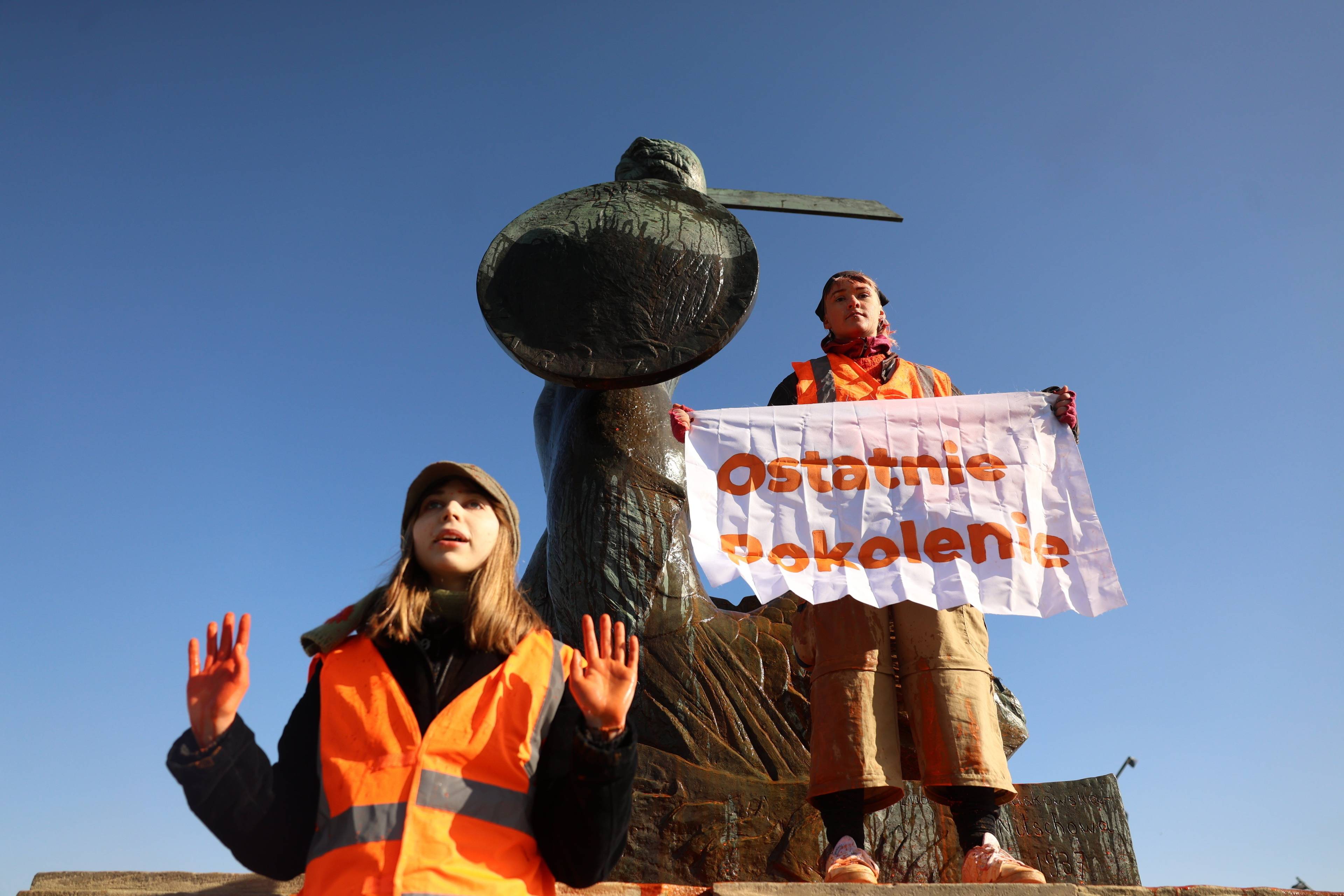 dwójka aktywistów, jedno z nich trzyma napis "ostatnie pokolenie", stoją na pomniku syrenki