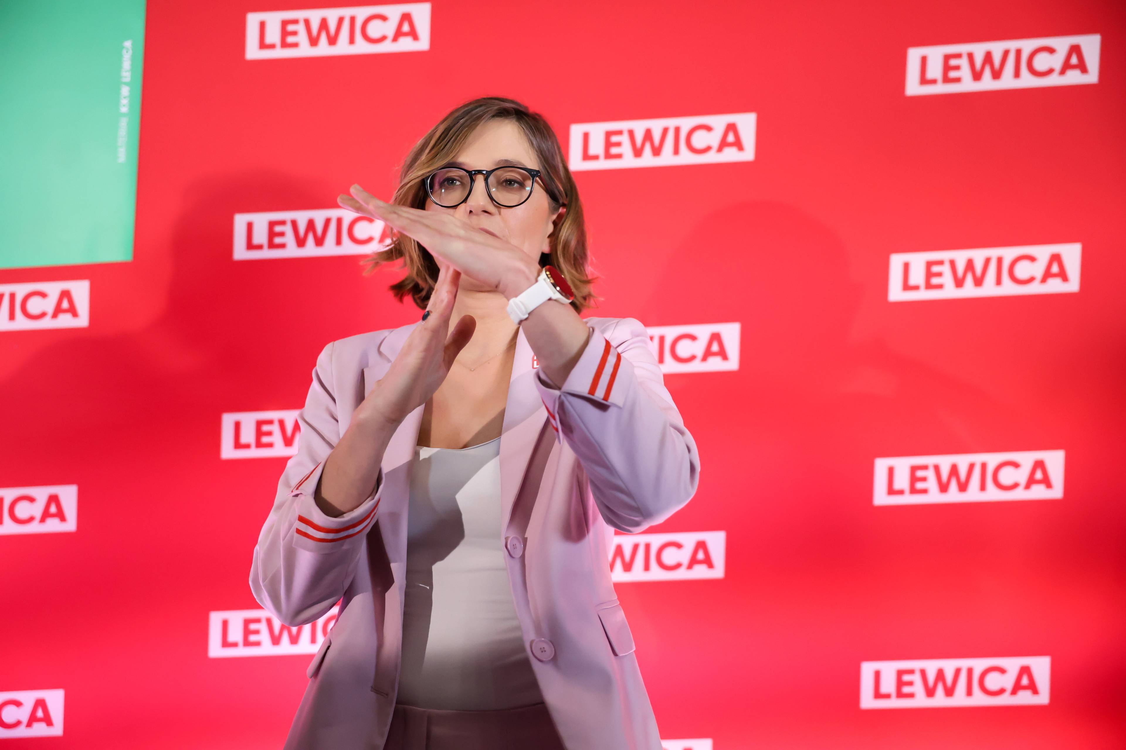 Młoda kobieta z włosami do ramion, w okularach (Biejat) stoi na tle czerwonej ścianki z logo Lewicy i pokazuje gest „czas się skończył”. Lewica"