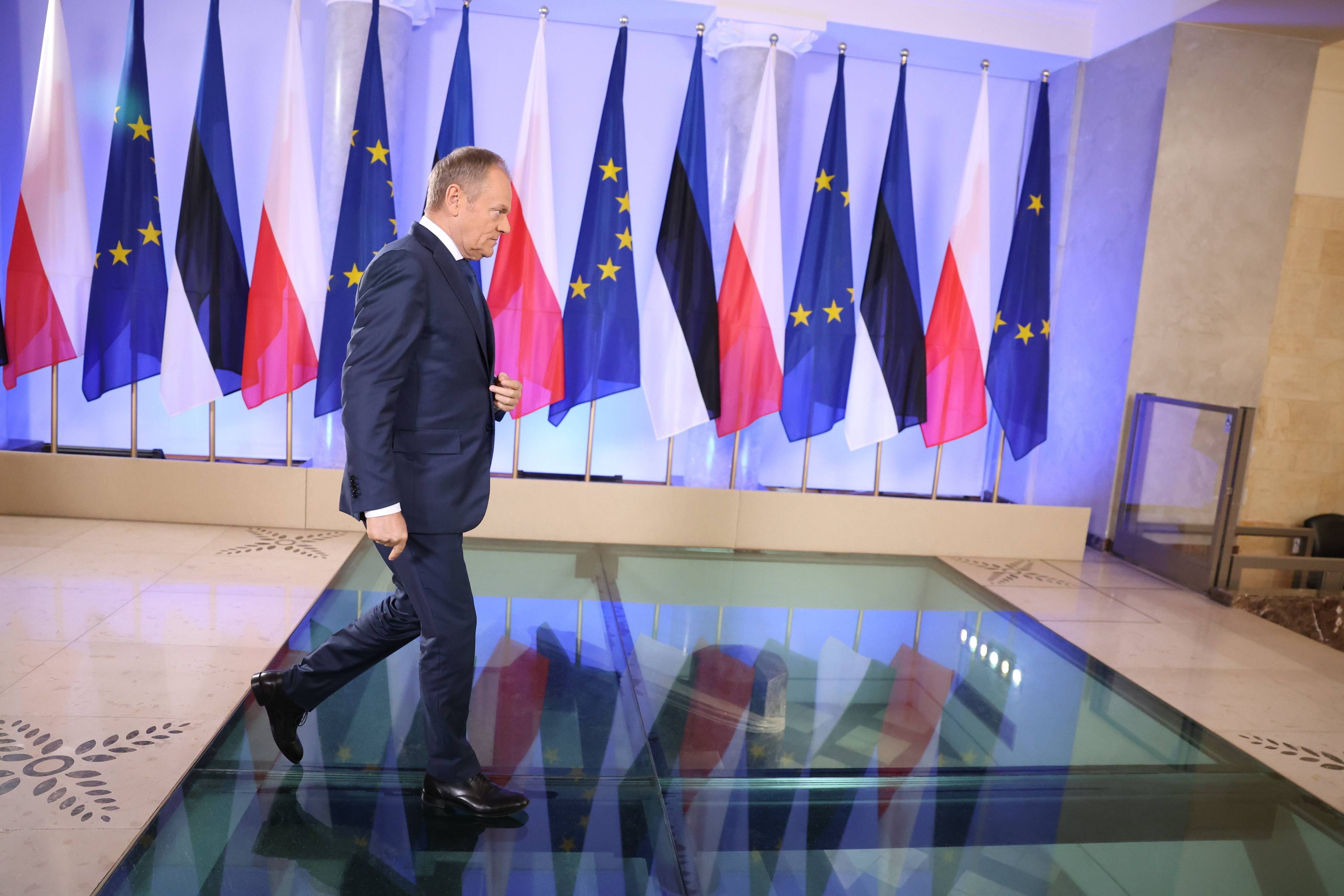 Donald Tusk przechodzi sam przez dużą salę, za nim ustawione w rzędzie flagi Polski i Unii Europejskiej