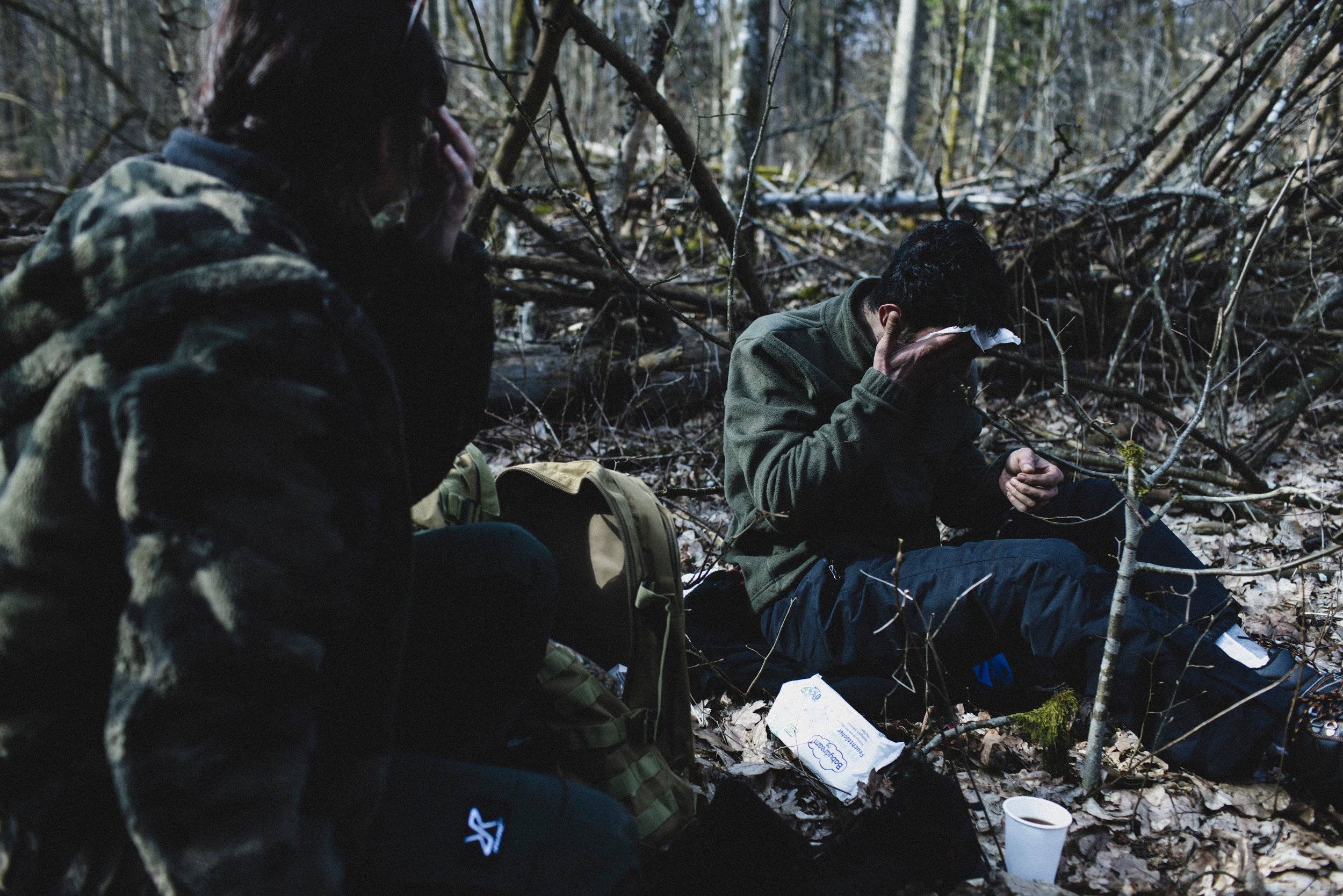 Dwaj uchodżcy siedzą w lesie, jeden z nich wyciera chusteczką higieniczną twarz, drugi okrywa się kocem. Granica