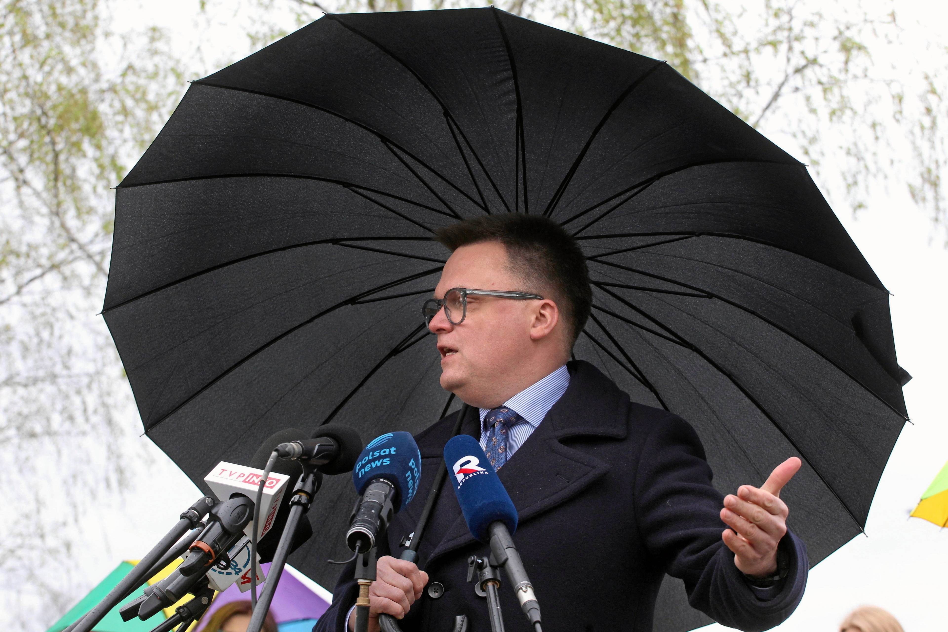 Szymon Hołownia trzyma parasol, przed nim pęk mikrofonów