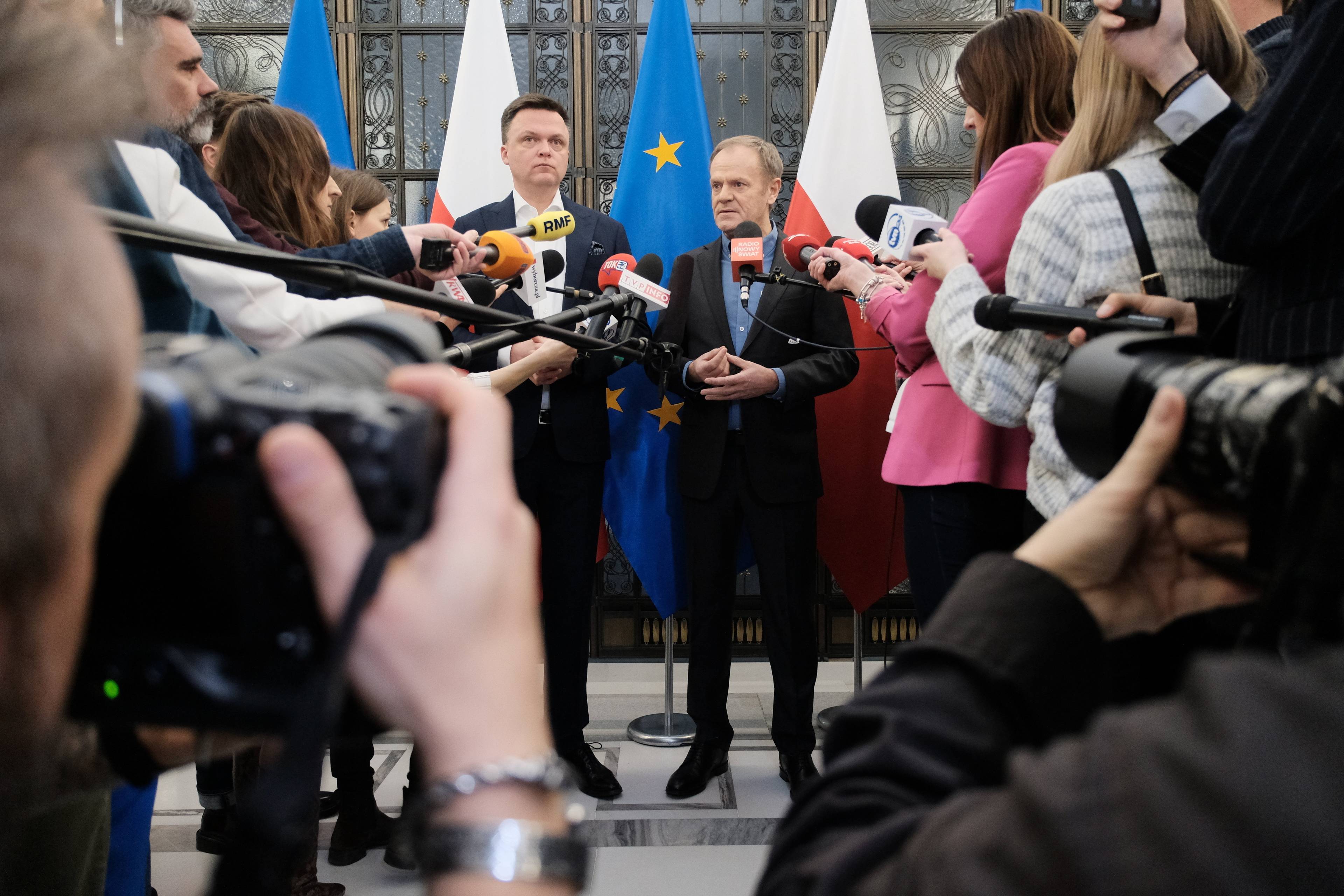 Donald Tusk i Szymon Hołownia stoją przed mikrofonami, kamerami a aparatami fotograficznymi dziennikarzy