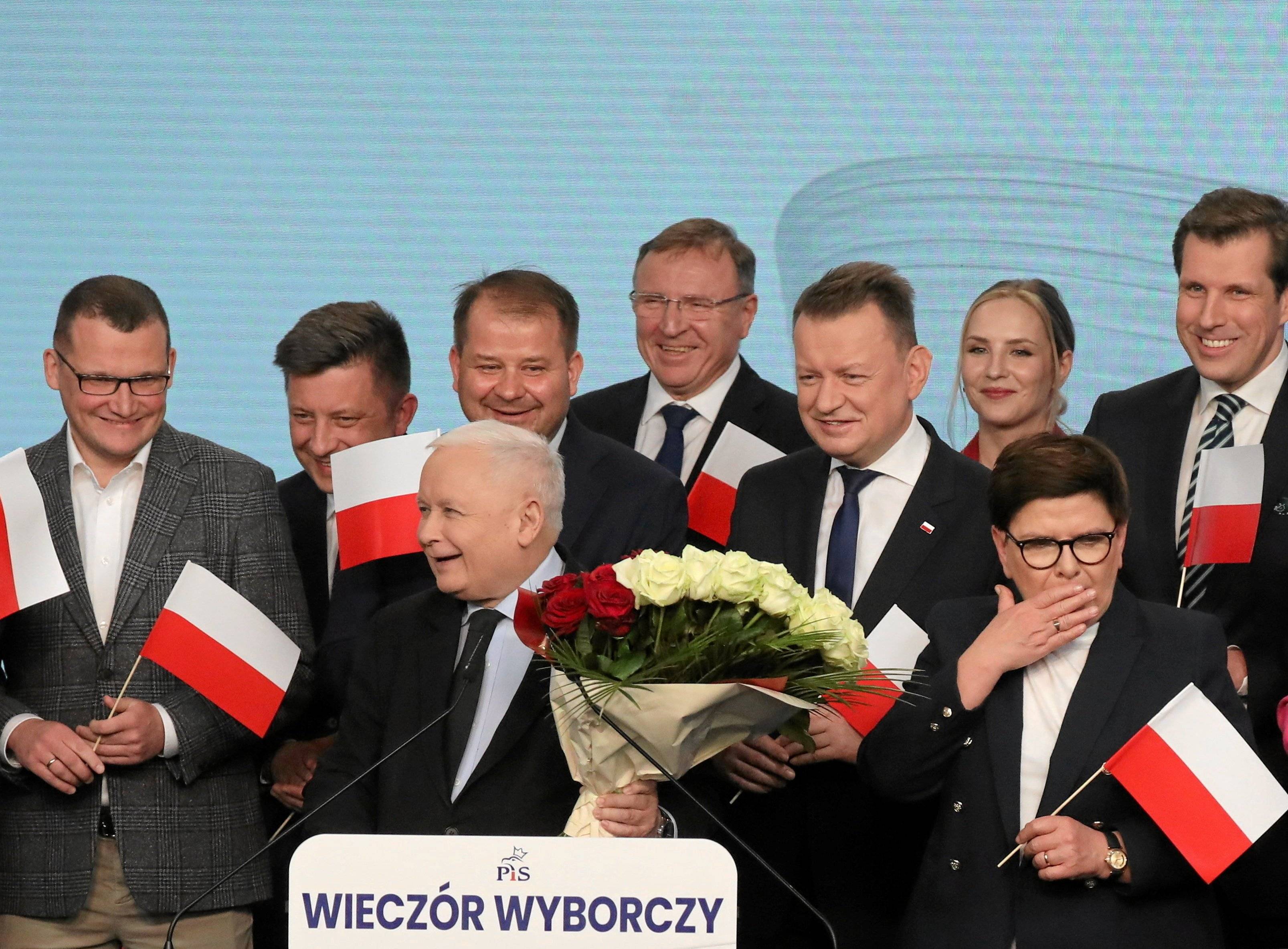 Rozradowany Jarosław Kaczyński - niski siwy starszy pan - w otoczeiu mężczyzn z biało-czerwonymi flagami, z biało-czerwonym bukietem w dłoni