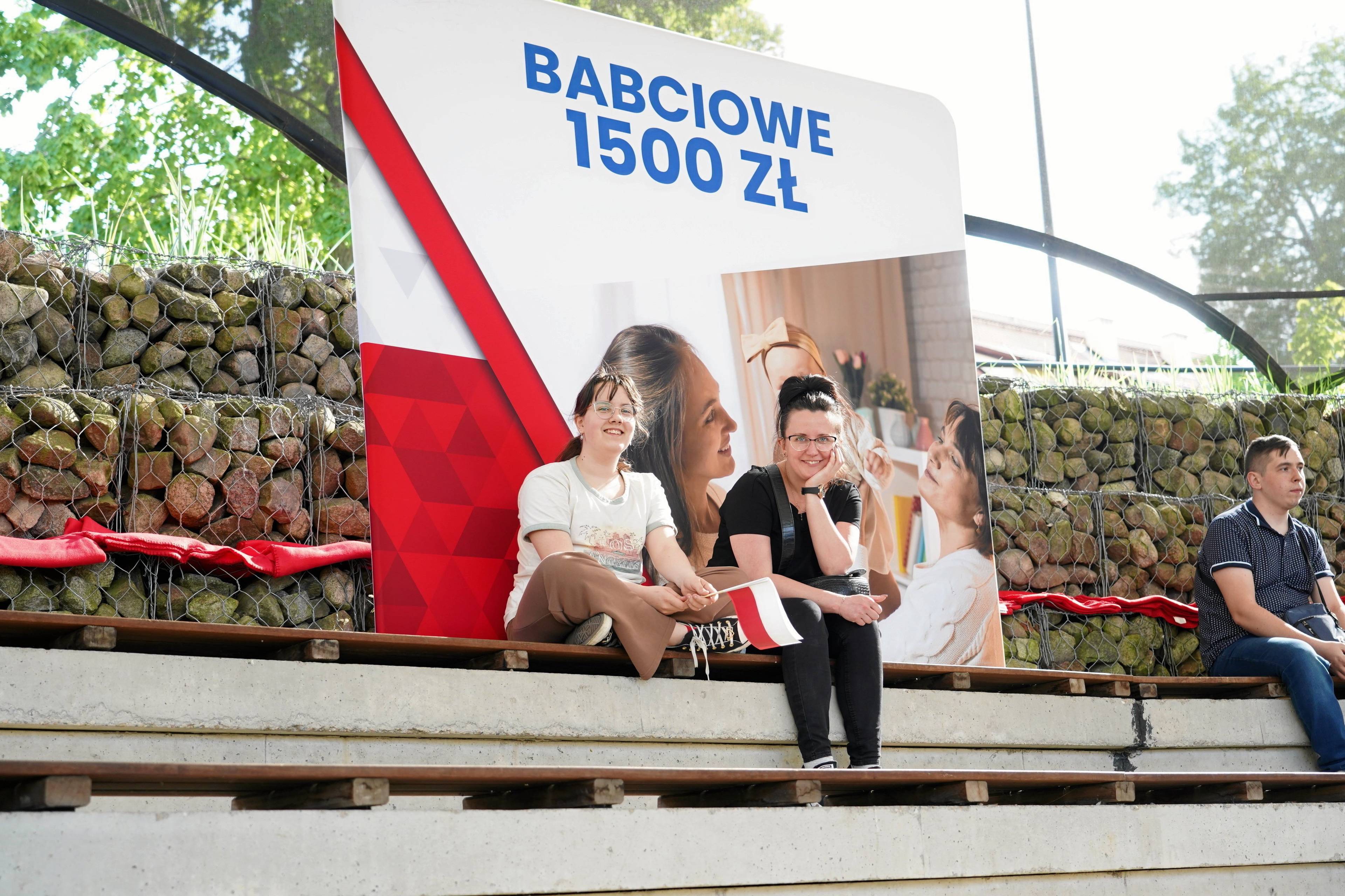 Dwie dziewczyny siedzą na tle plakatu z transparentem "Babciowe 1500 zł"